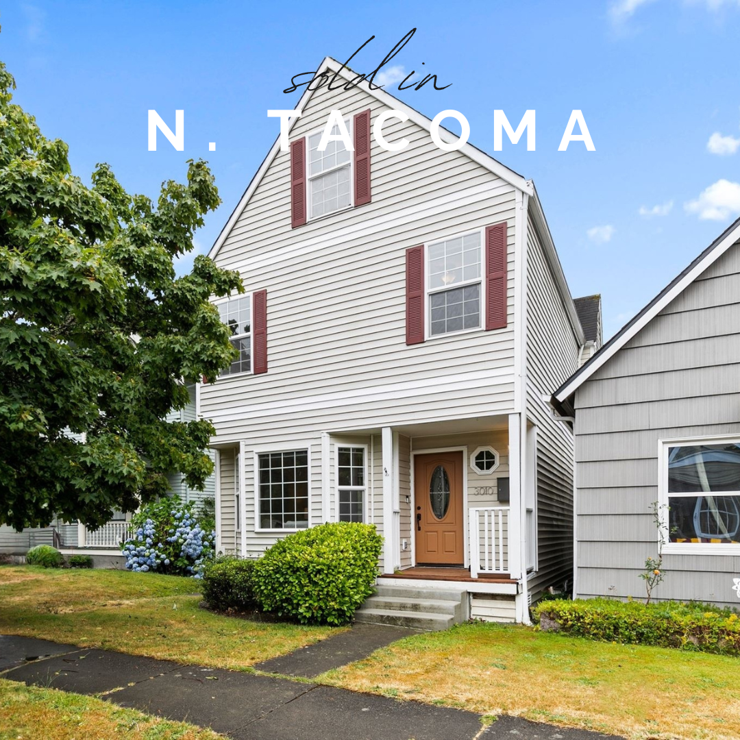 White multi-story house with red shutters, a small front porch, and a brown front door, located in Tacoma, Washington, with green bushes and a tree in the yard.
