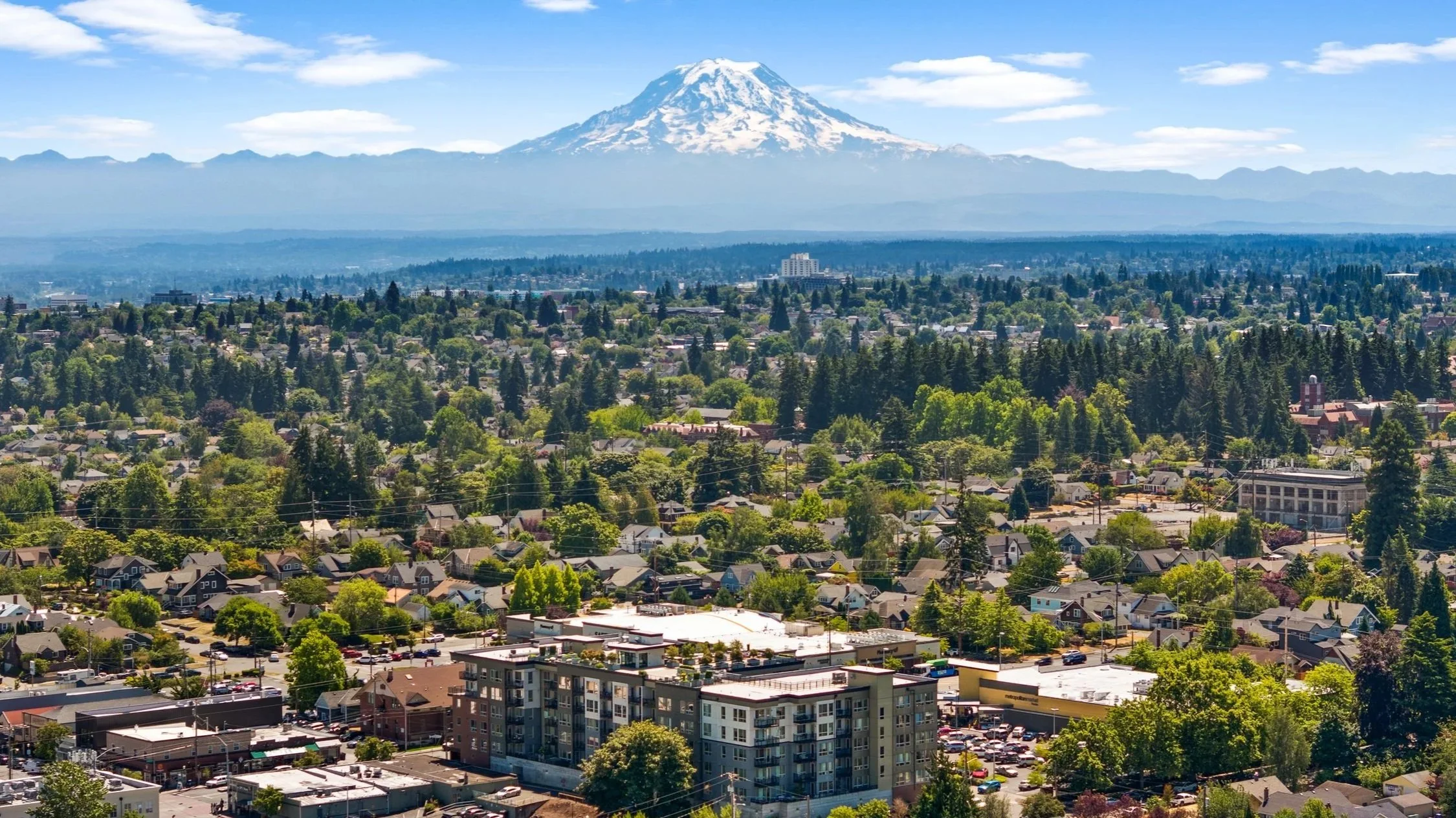 A cityscape with numerous houses, buildings, and trees, with Mount Rainier snow-capped in the background under a blue sky with light clouds.