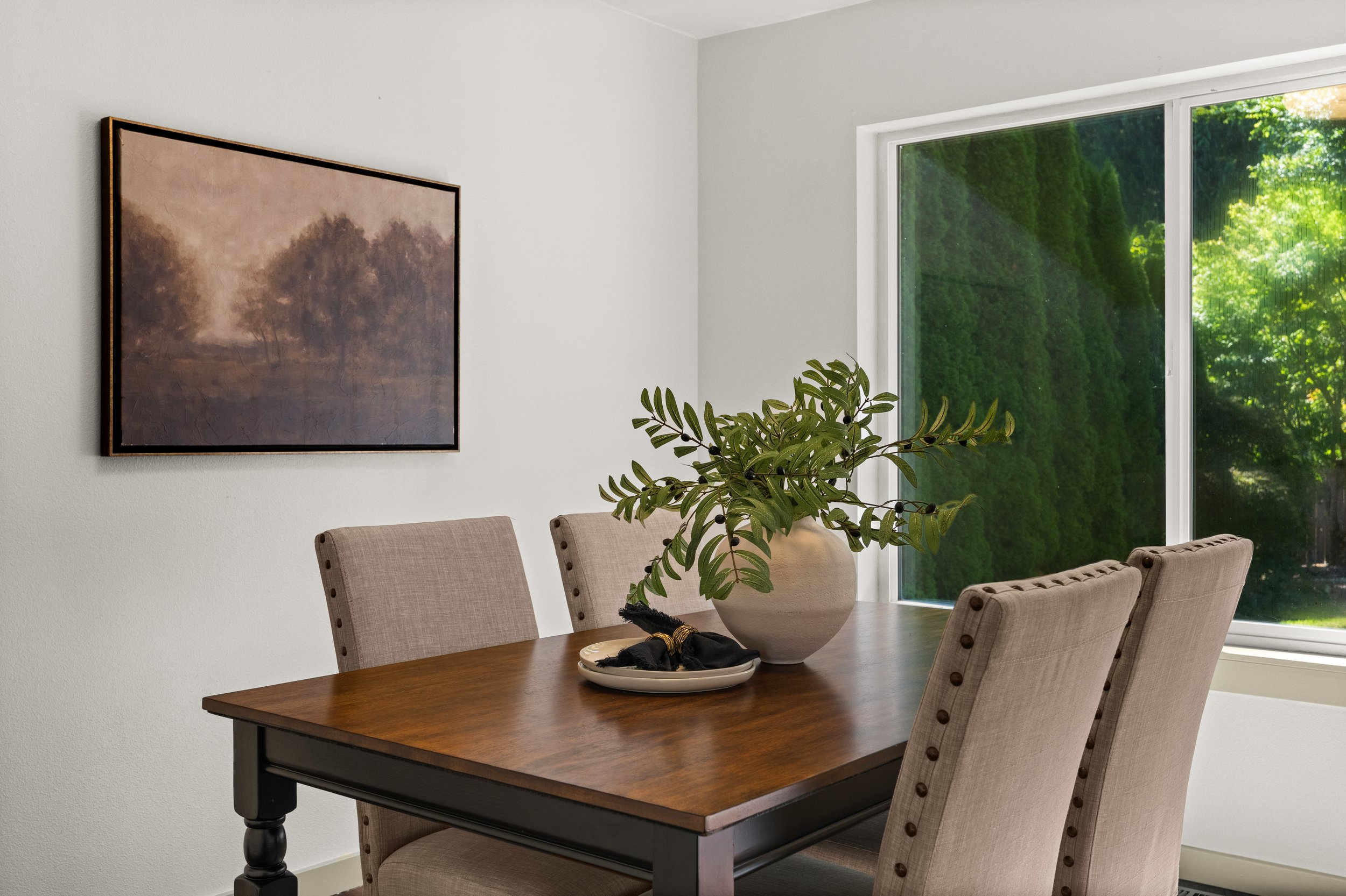 A dining room with a wooden table surrounded by beige upholstered chairs. A large potted plant with green leaves is on the table. A window shows lush green trees outside, and a framed landscape painting hangs on the white wall.