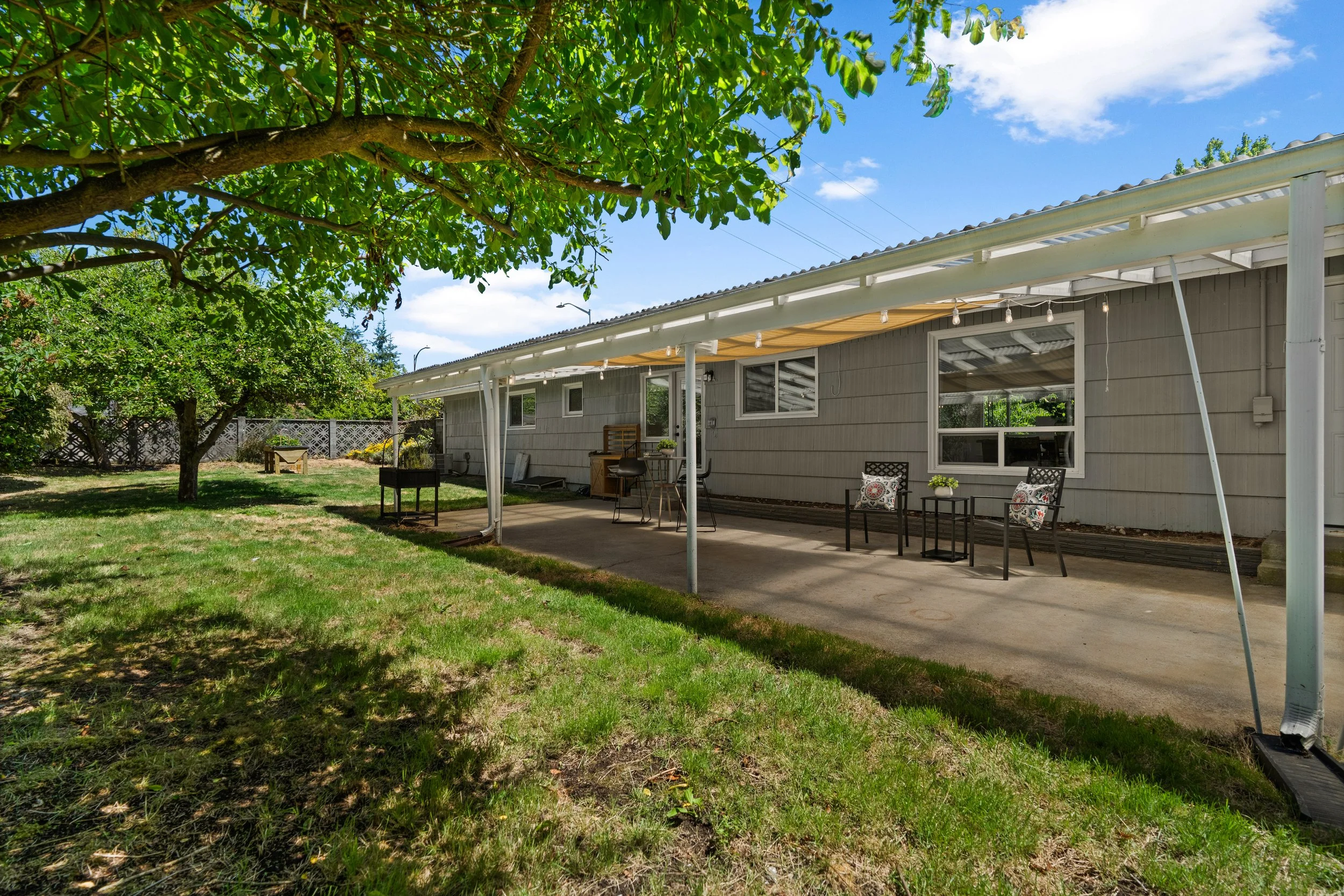 Backyard patio area with a house, table, chairs, and string lights under a large tree on a sunny day.
