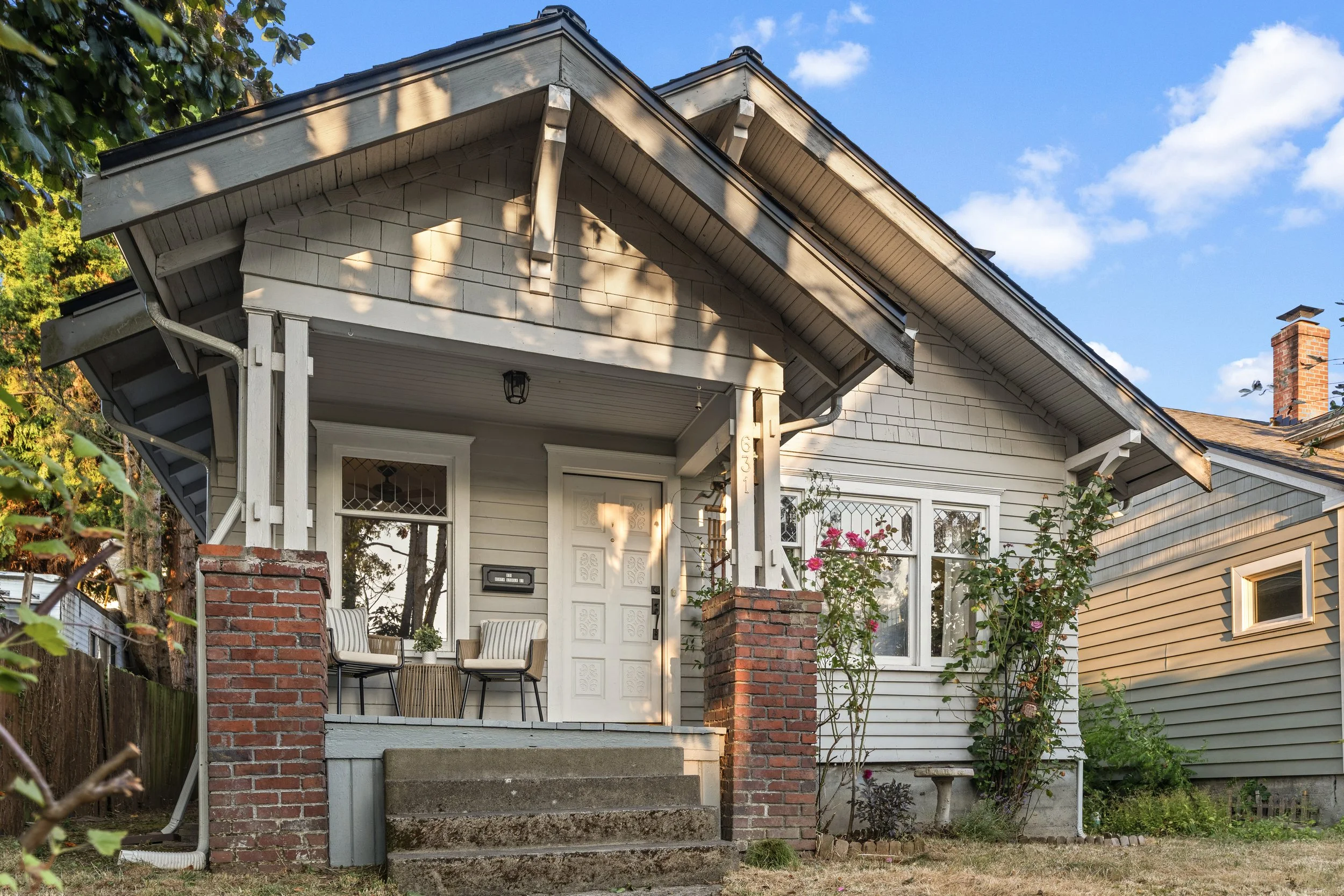 Front porch of a house with brick steps, white wooden columns, a white door, and a seating area with striped chairs. The house has gray siding, a gabled roof, and a brick chimney, and is surrounded by trees and plants under a blue sky with some clouds.