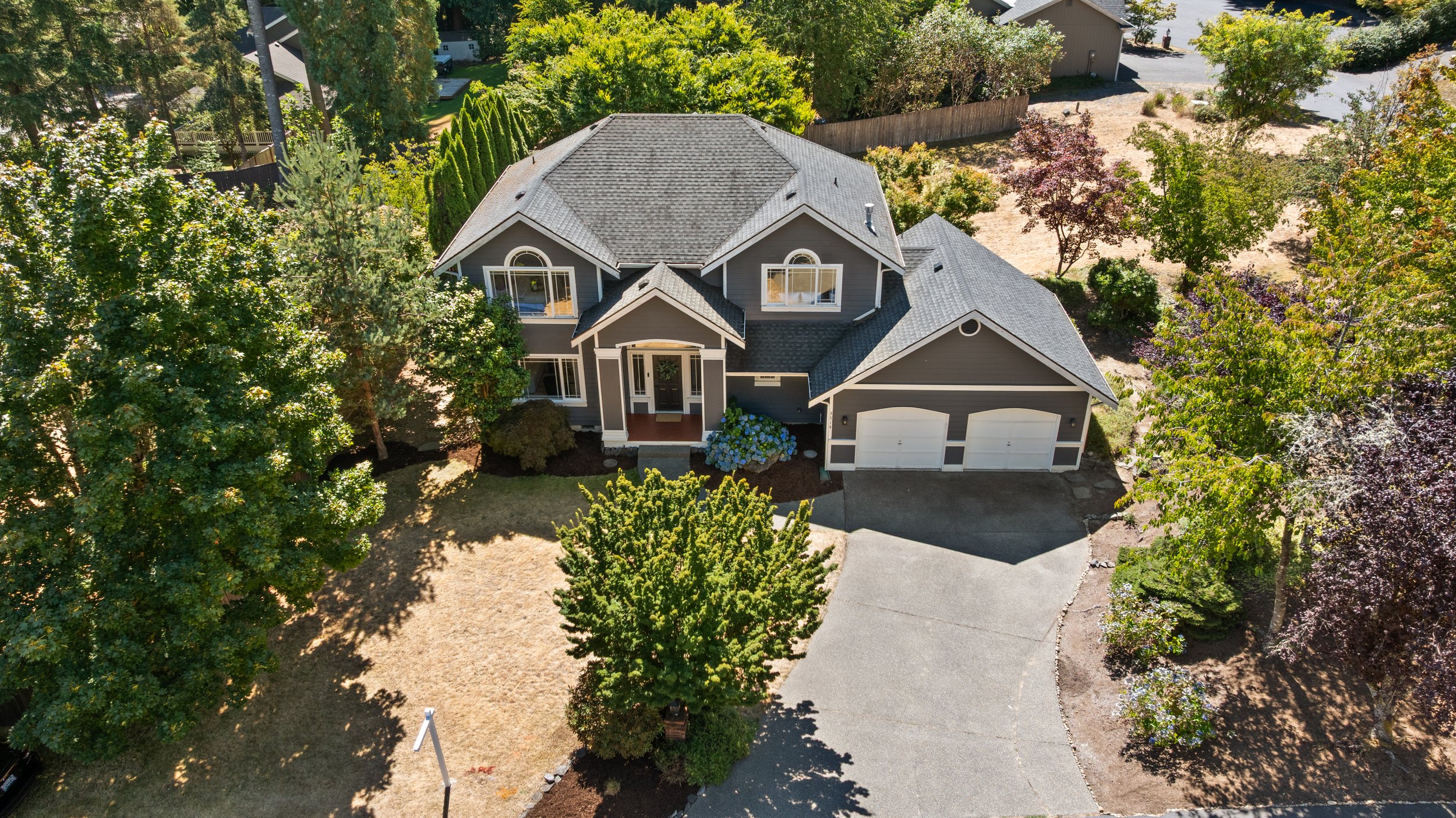Aerial view of a two-story suburban house with gray siding, a gabled roof, and a two-car garage, surrounded by green trees and landscaped yard.
