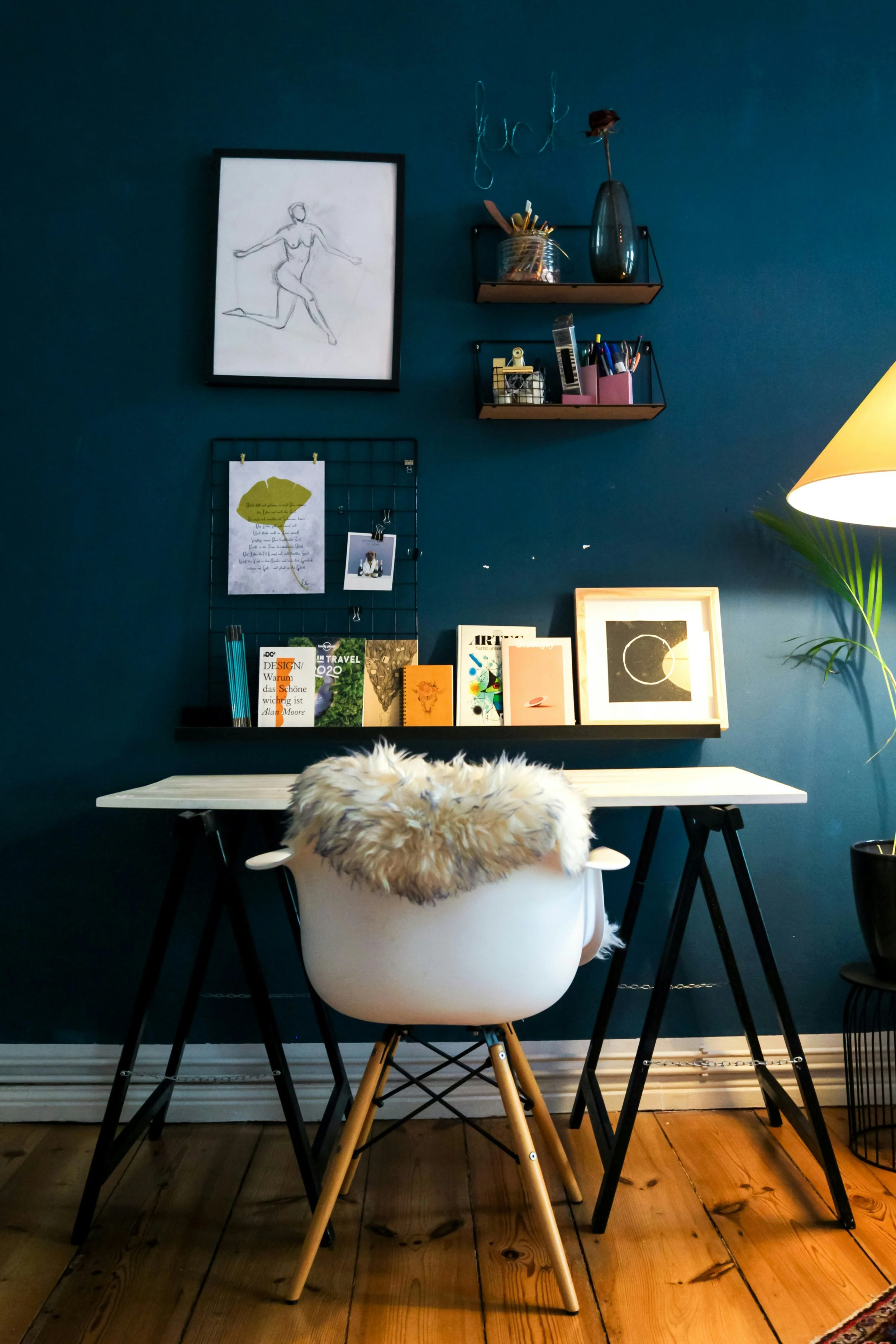 A cozy home office with a white desk and a white chair draped with a wool throw. The wall behind is painted dark blue and decorated with artwork, books, and shelves holding desk supplies. A potted plant is partially visible on the right with a yellow lampshade nearby.