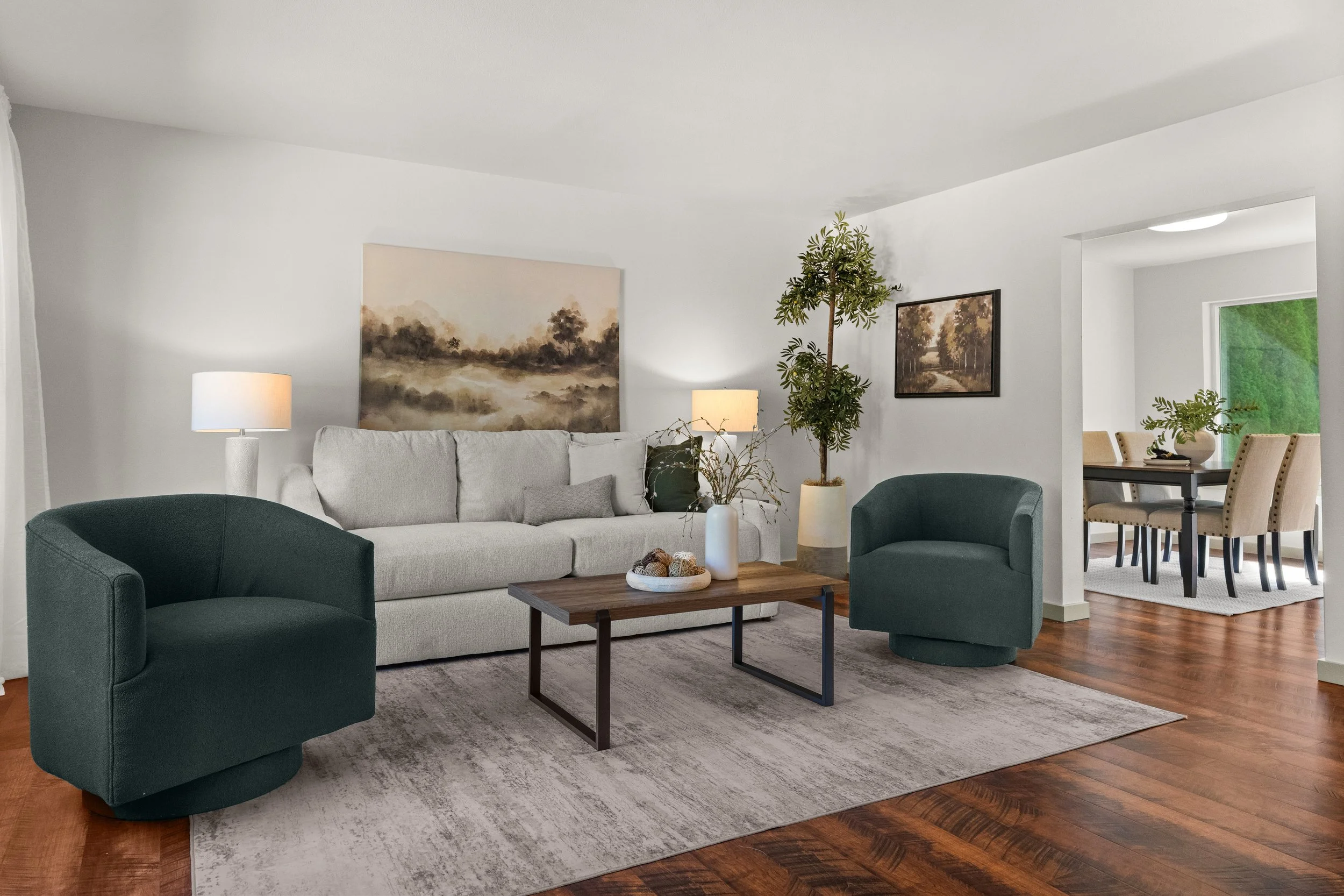 Living room with a white couch, two green armchairs, a wooden coffee table, and wall art depicting nature scenes. There are two lamps and a large potted plant. A dining area with a table and beige chairs is visible in the background.