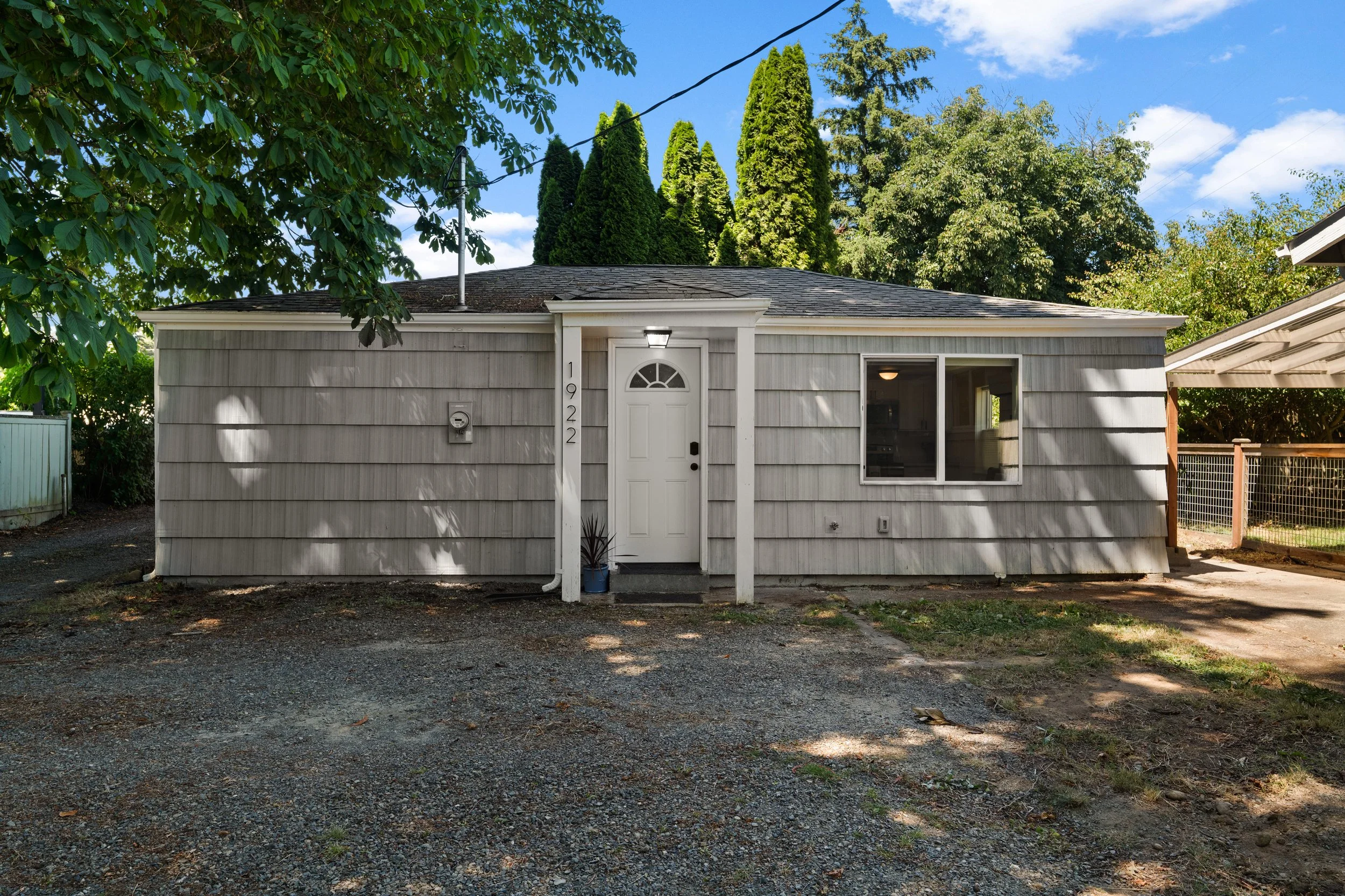 A single-story house with white horizontal siding, a white door with an arched window, and a small porch area. The house has a gray shingle roof, and there is a blue plant pot near the entrance. Surrounding trees and blue sky with some clouds are visible in the background.