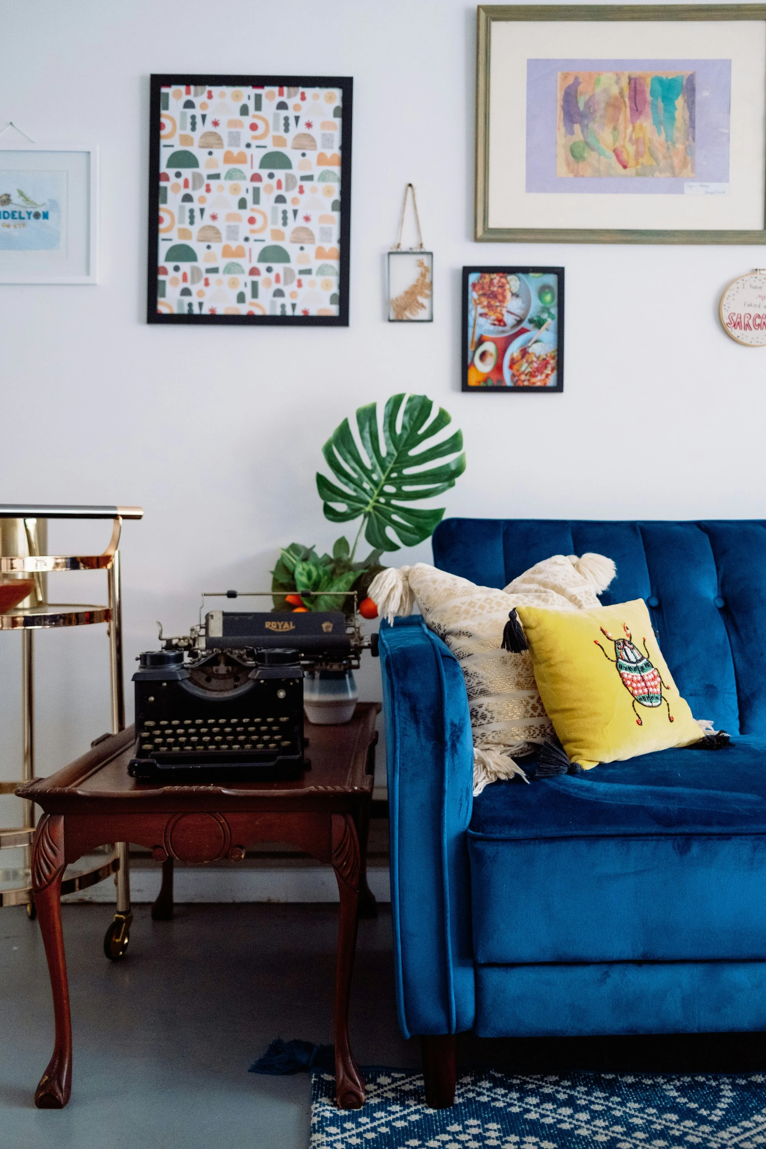 Living room with a blue velvet sofa, decorative pillows, vintage typewriter on a wooden table, wall art including framed pictures and watercolor painting, large green monstera leaf, and a gold bar cart.