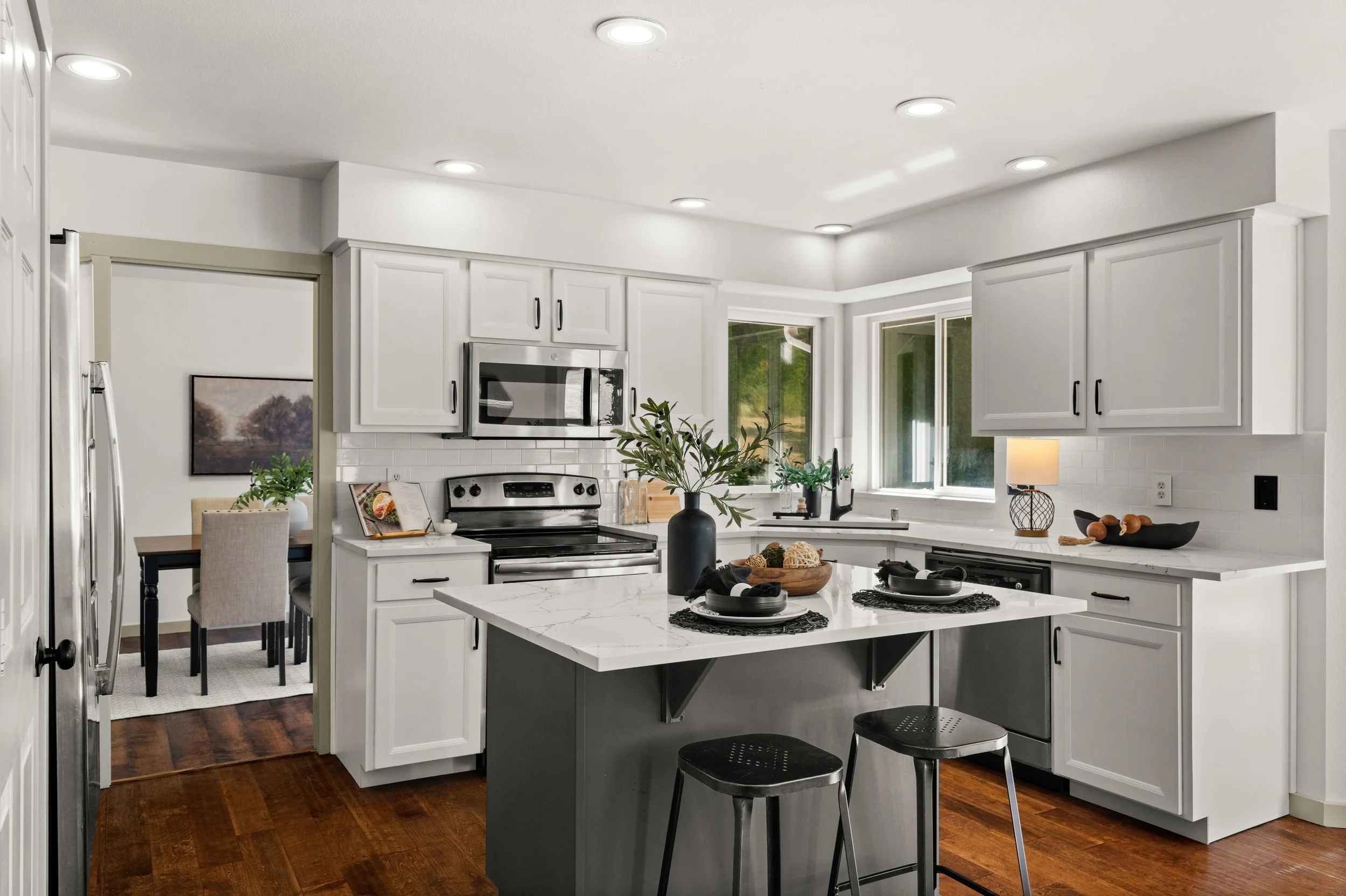 A modern kitchen with white cabinets, black hardware, and a white marble island. There are stainless steel appliances, including a microwave and stove. The kitchen has large windows, potted plants, and a dining area in the background.
