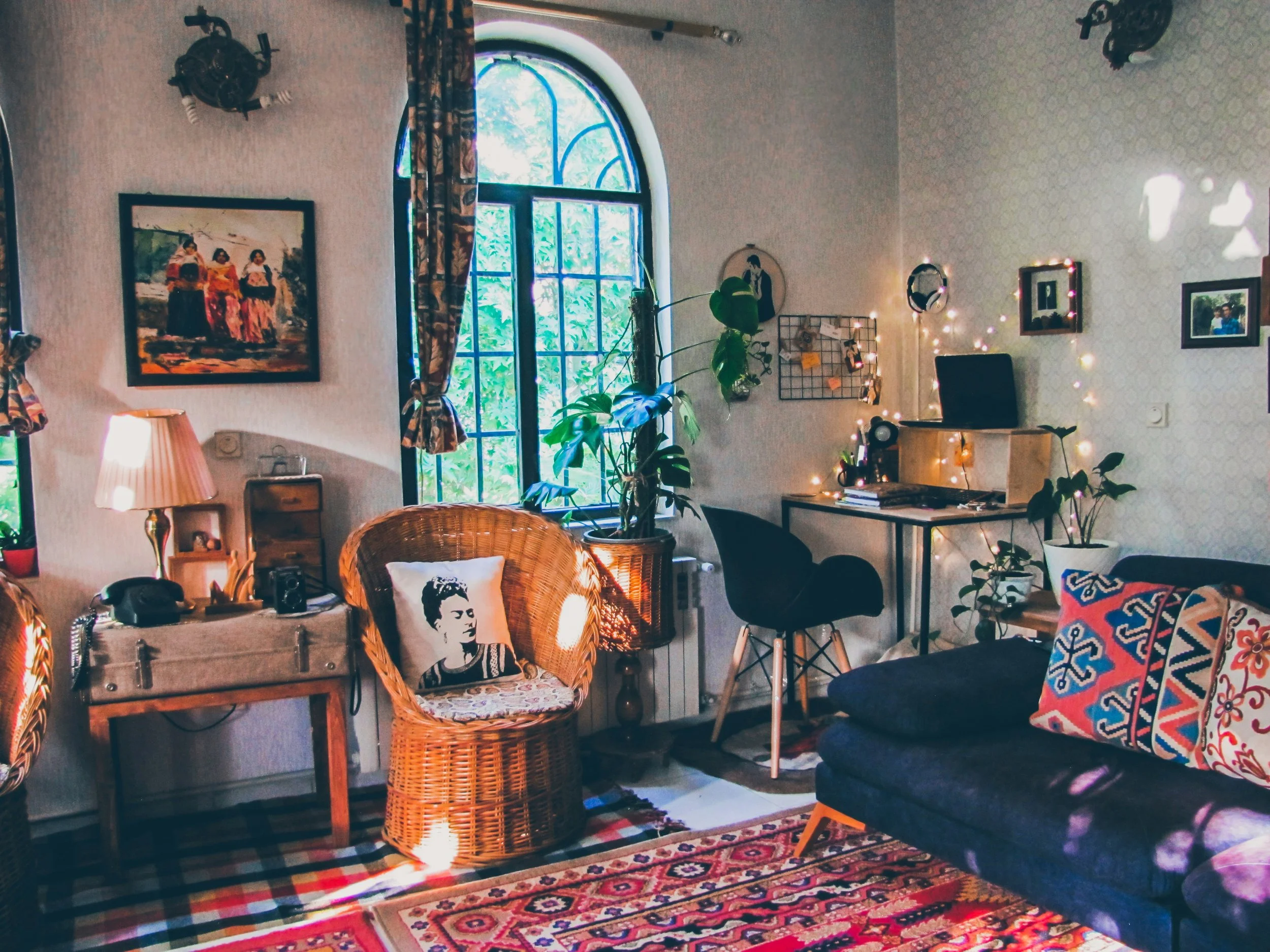 Cozy living room with a large window, a wicker chair with Frida Kahlo pillow, a black sofa with colorful pillow, a wooden desk with a computer, plants, artwork, and string lights.