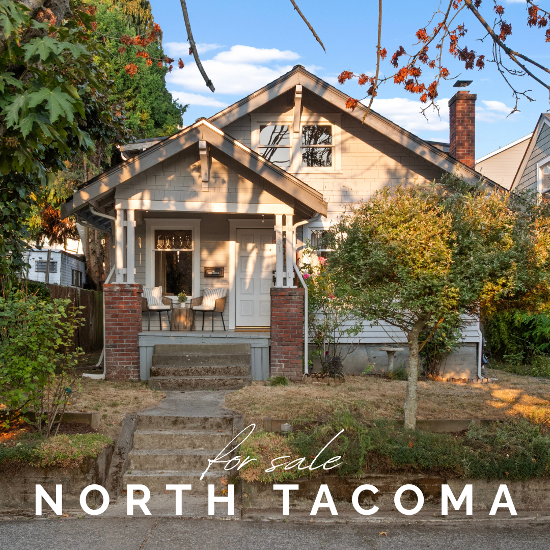 A house with a small front porch, two chairs, and steps leading up to the entrance. Trees surround the house, with a "for sale" sign on the lawn, in North Tacoma.