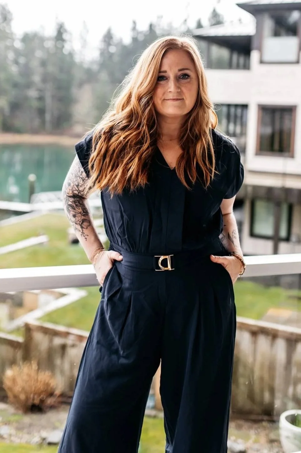 A woman with long wavy red hair, wearing a black jumpsuit with a belt, standing on a balcony with a view of a lake, trees, and modern apartment buildings in the background.