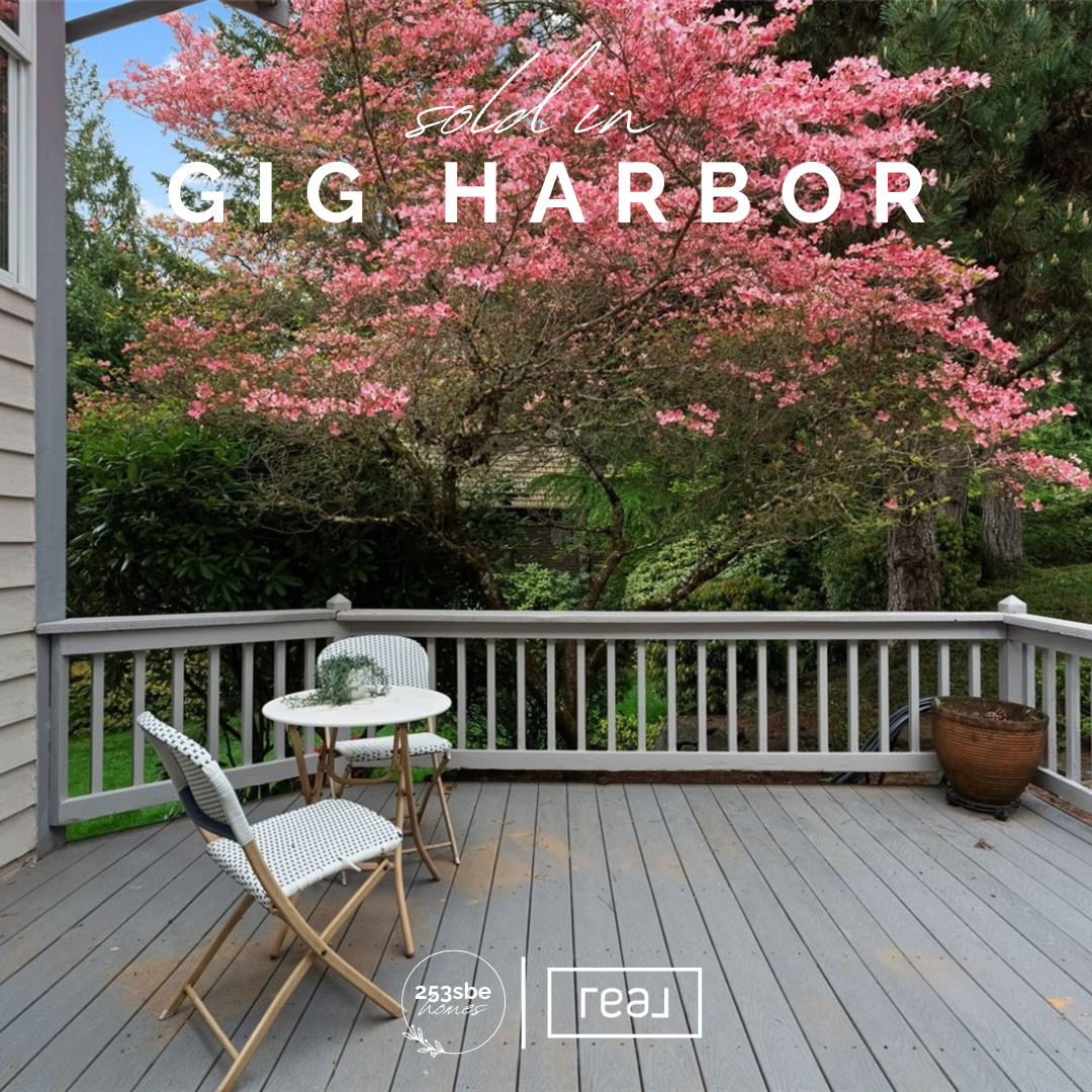 A porch with wooden decking, white railing, a round table with two chairs, a large potted plant, and a blooming pink tree in the background.