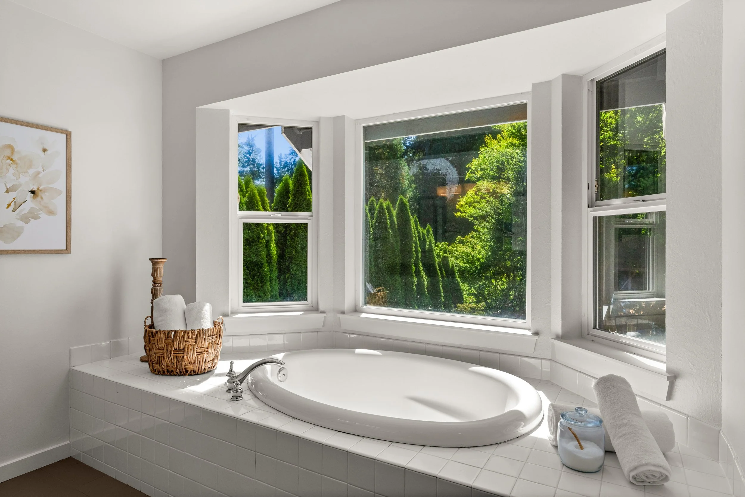 A bright bathroom with a white bathtub surrounded by white tiles and three large windows showing lush green trees outside. There is a wicker basket with rolled white towels on the tub's edge, a glass container, and a towel rolled up next to it.