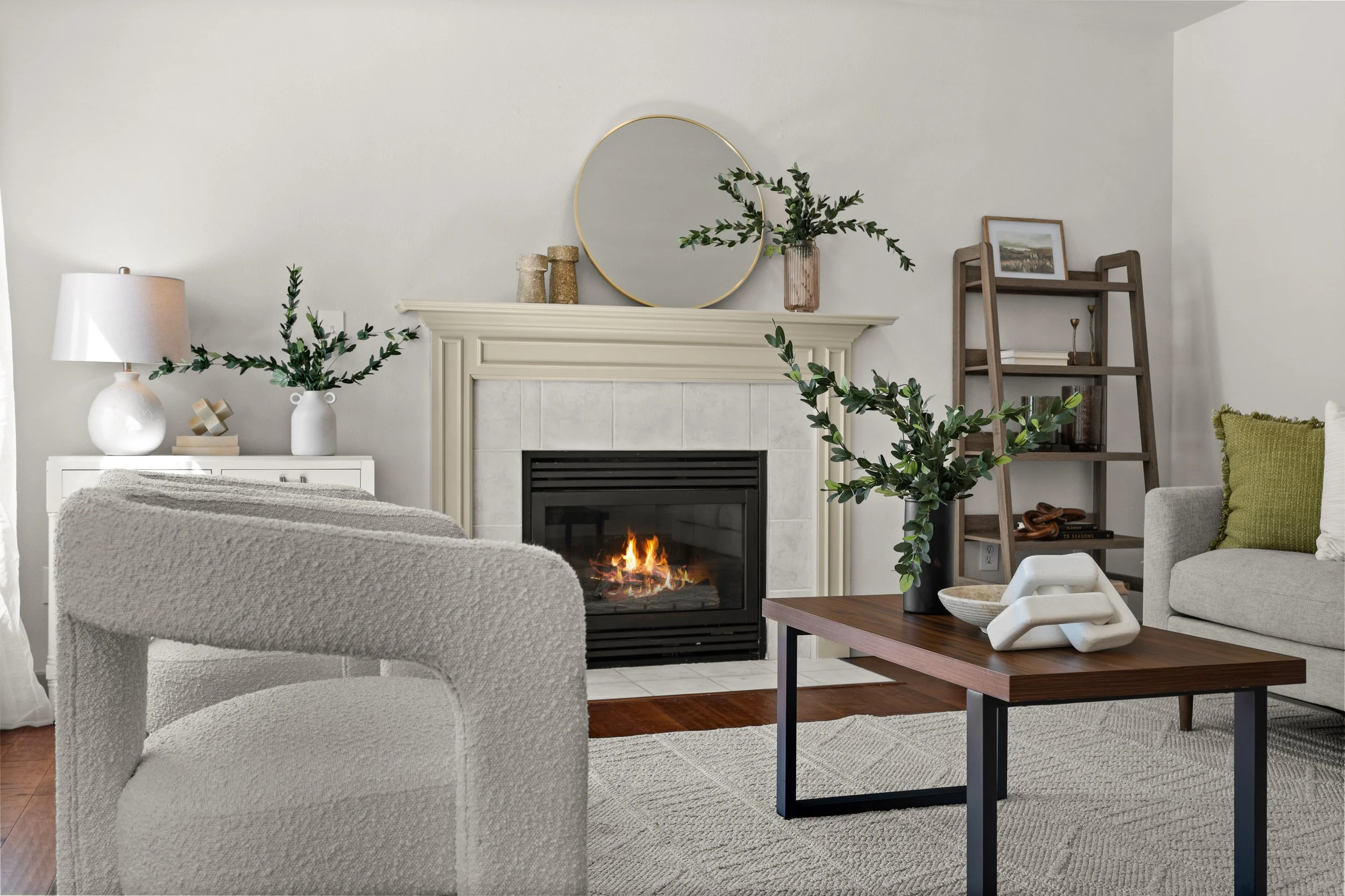 A cozy living room with a fireplace, beige armchair, coffee table, and wooden shelving. Decor includes vases with greenery, a mirror above the fireplace, framed artwork, and candles.