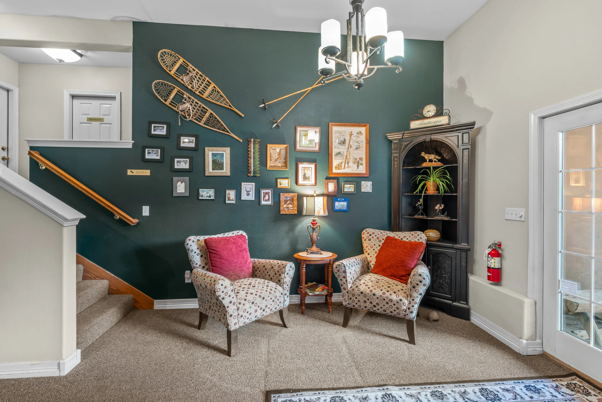 Living room corner with two patterned armchairs, coral pillows, a small round side table with a lamp, a dark wooden cabinet with a plant on top, collage of framed pictures and vintage skis on a teal wall, chandelier, and glass door to the right.