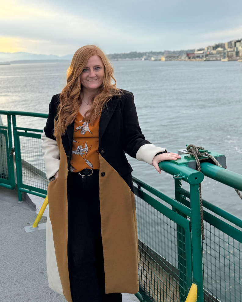 A woman with long red hair smiling, standing on a boat deck near water, with a city and hills in the background.