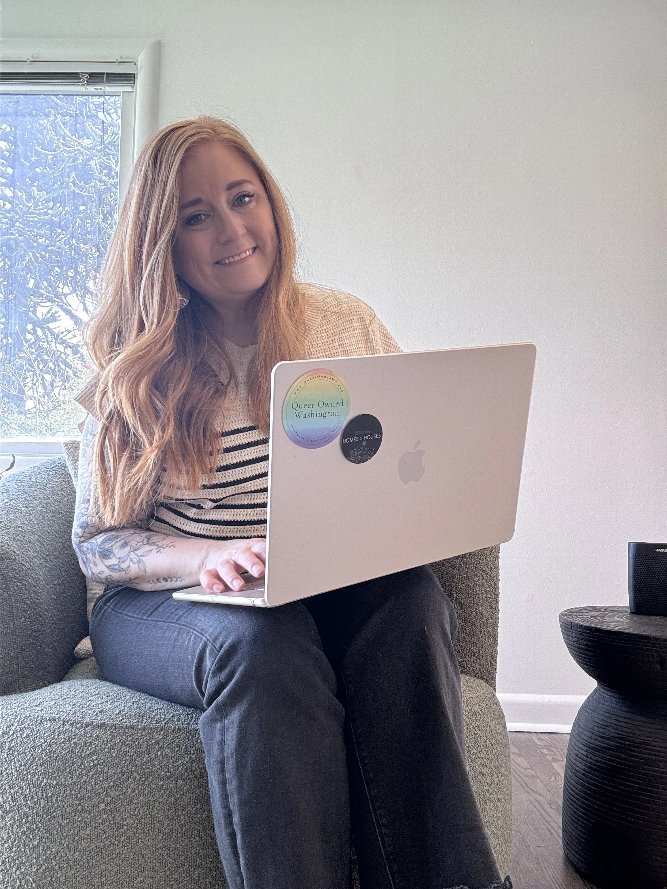 A woman with long red hair sitting on a couch, smiling at the camera, using a silver Apple MacBook with stickers on it, in a room with a window showing snow outside.