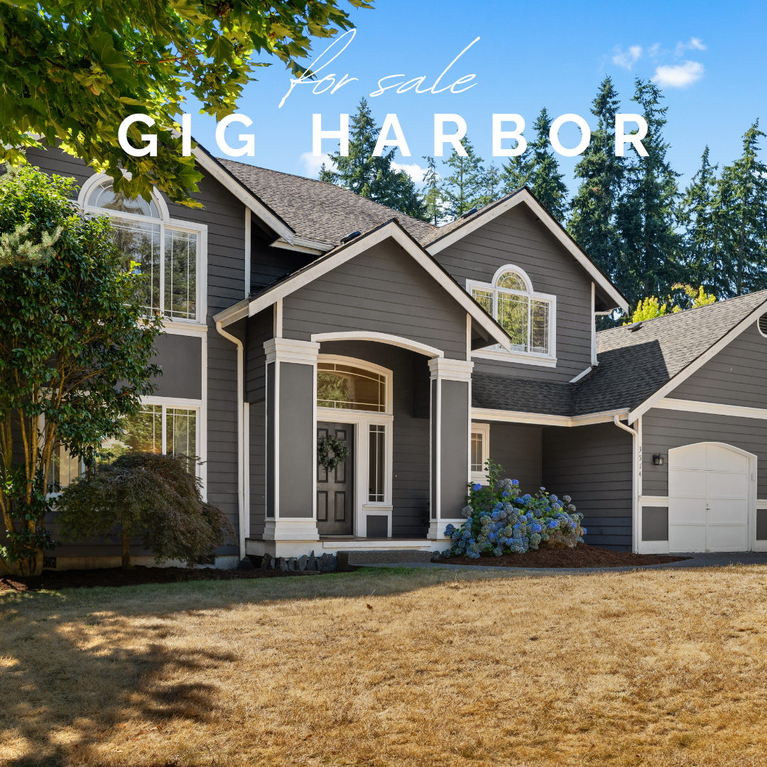 Front view of a gray house with white trim, surrounded by trees and bushes, with a lawn in the foreground. The house has large windows and a front door with a wreath. Text overlay says 'For Sale GIG HARBOR'.