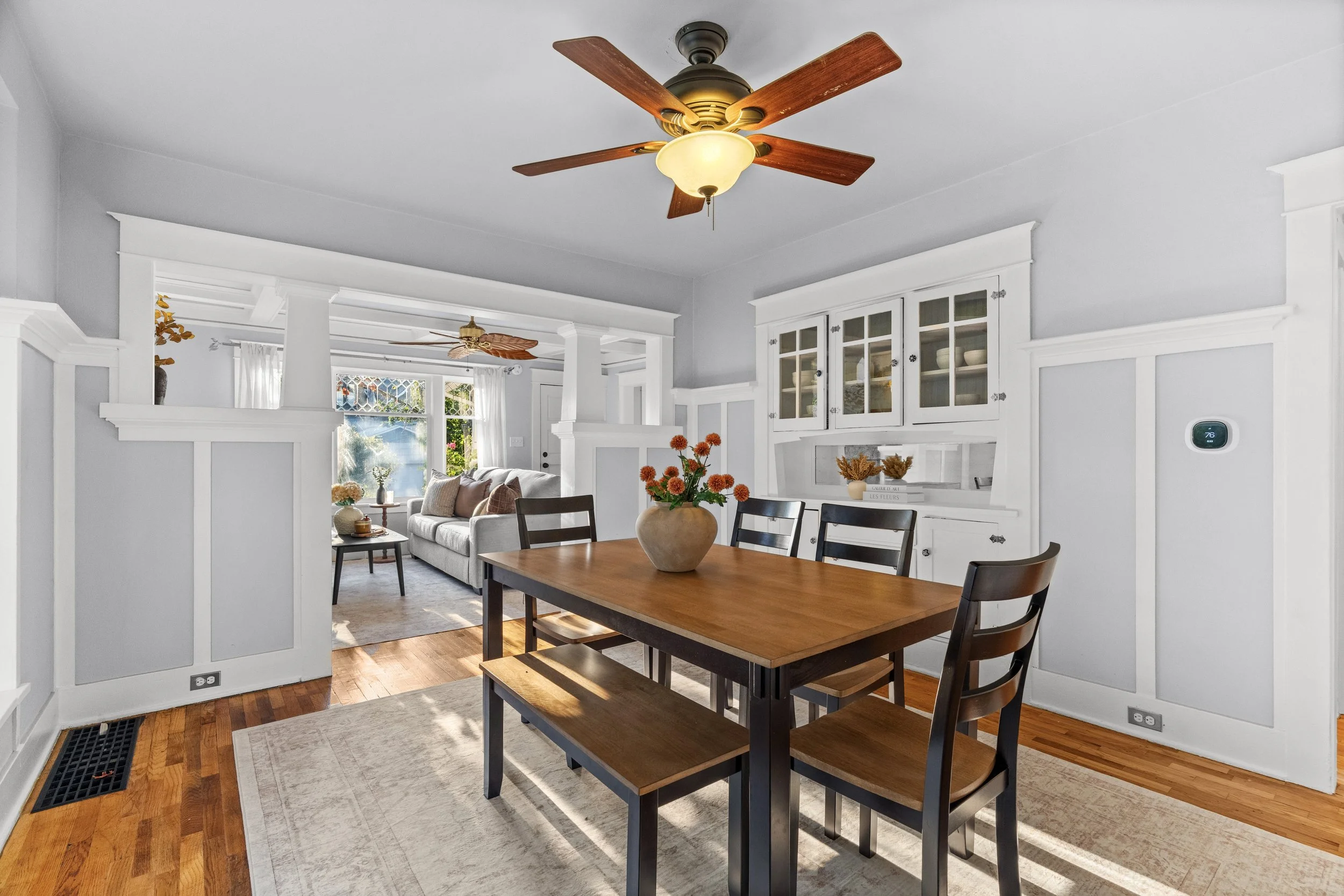 Open-concept living and dining room with white walls, wooden flooring, a ceiling fan, a dining table with chairs, a sofa, and a window with curtains.