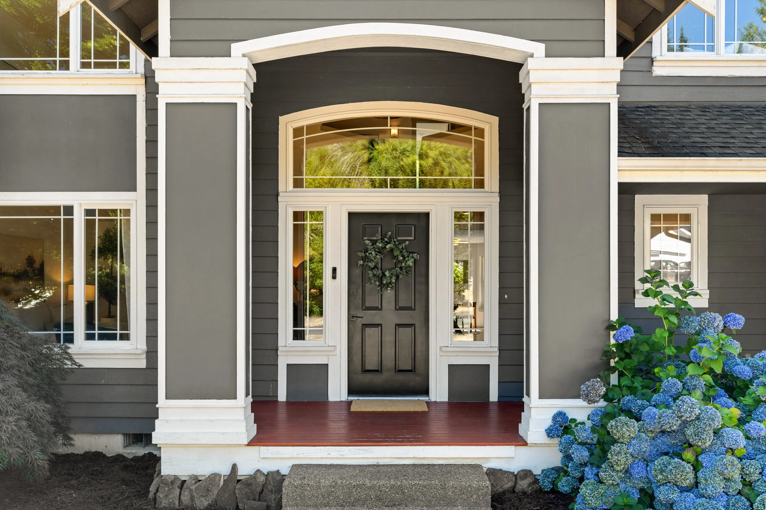 Front entrance of a modern gray house with white trim, featuring a black door decorated with a wreath, large windows, and a porch with red flooring. Blue hydrangeas are visible on the right side of the porch.