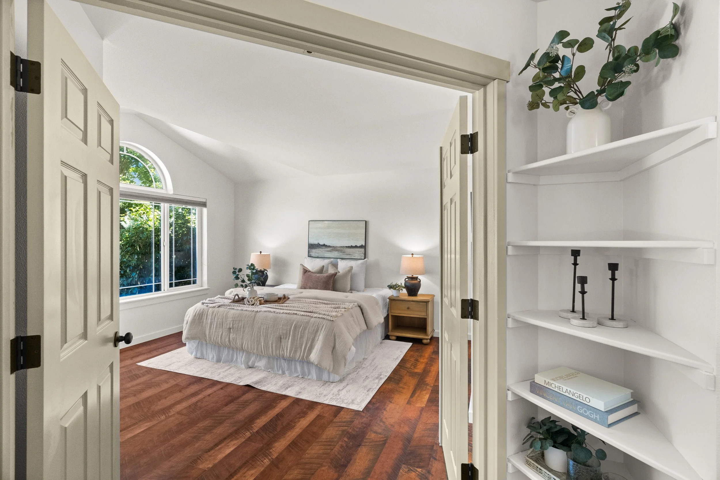 A cozy bedroom with a white bed, beige bedding, and pillows, facing a large window with an arched top, displaying green trees outside. There are two nightstands with lamps, and a framed artwork hangs above the bed. The room has hardwood floors and a white wall opposite the bed. A white shelf with books and decorative items is visible on the right side of the image, seen through an open white door.