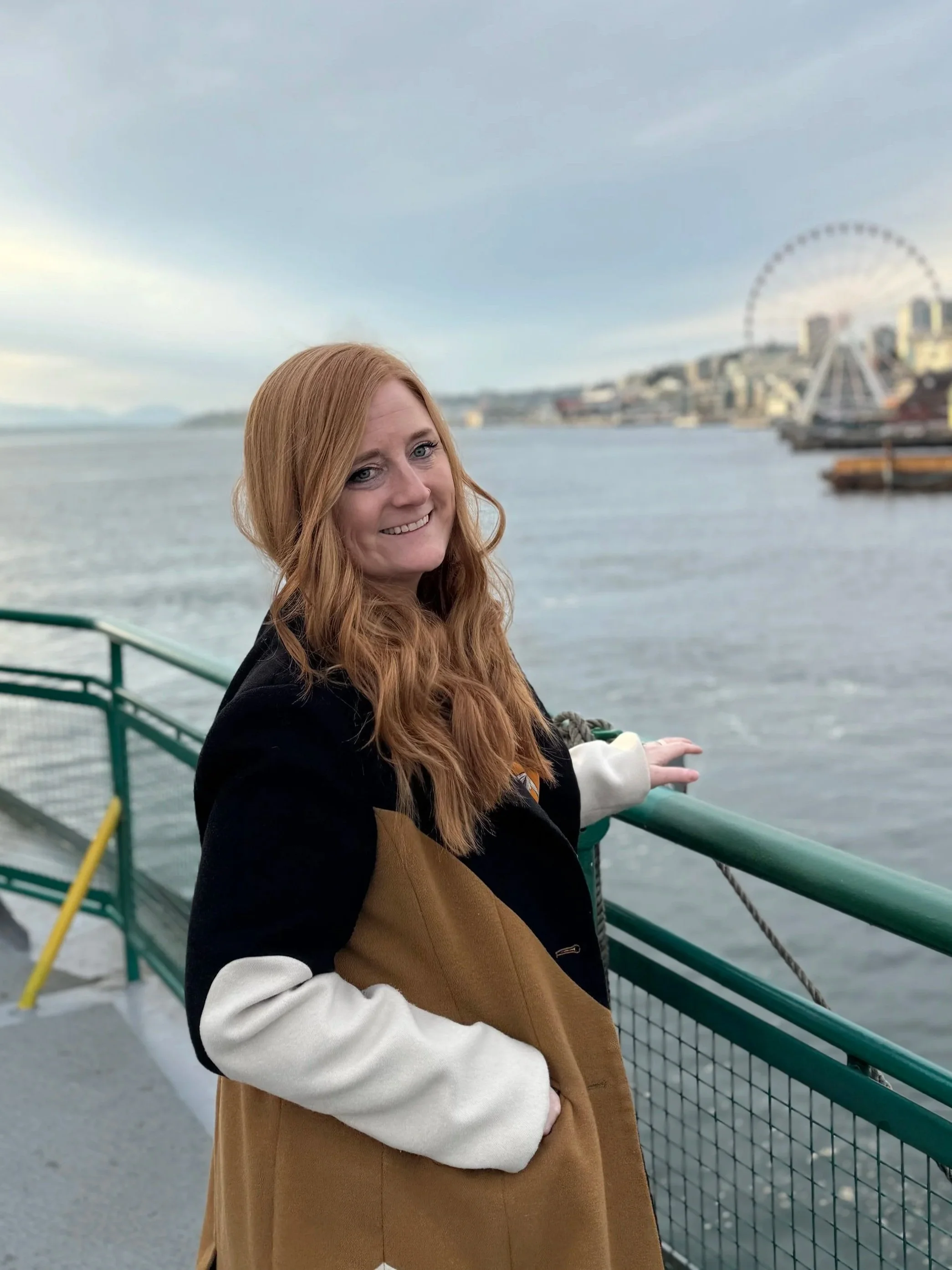 A woman with red hair smiling on a boat or ferry, with a waterfront cityscape, a Ferris wheel, and a cloudy sky in the background.