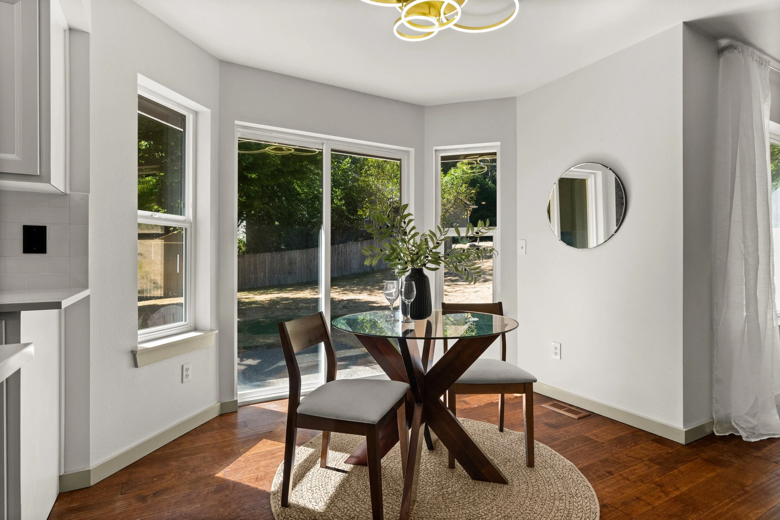 A dining area with a round glass table, two wooden chairs with beige cushions, a black vase with green foliage, and two wine glasses. Large windows with white frames let in natural light and show green trees outside. A round mirror hangs on the white wall, and white curtains are on the right. The floor is hardwood with a small beige rug under the table.