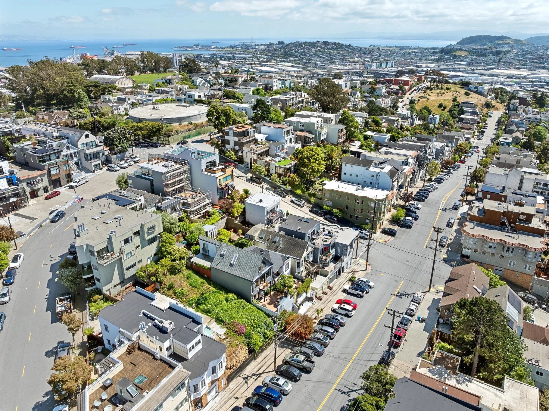 Aerial view of a residential neighborhood near a harbor with ships, industrial buildings, and water in the background. The neighborhood features a mix of modern and older houses, streets lined with parked cars, trees, and green spaces.