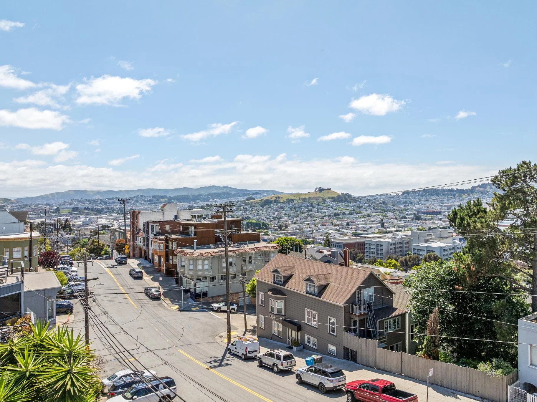 A cityscape with residential and commercial buildings, hills in the background, and a bright blue sky with clouds.