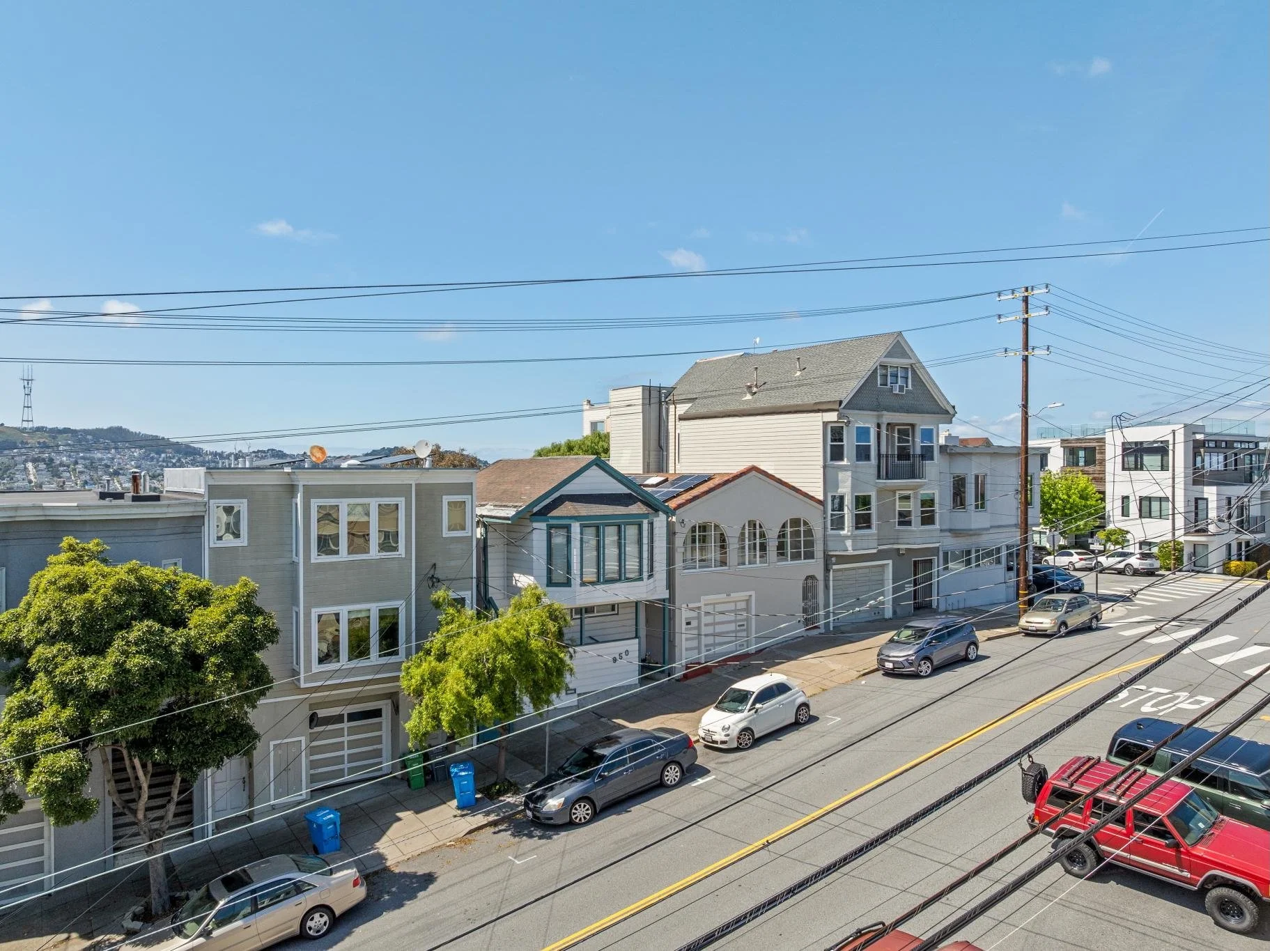 A residential street view with several multi-story houses, parked cars along the street, utility poles with multiple wires, and some greenery in front of the houses, under a clear blue sky.
