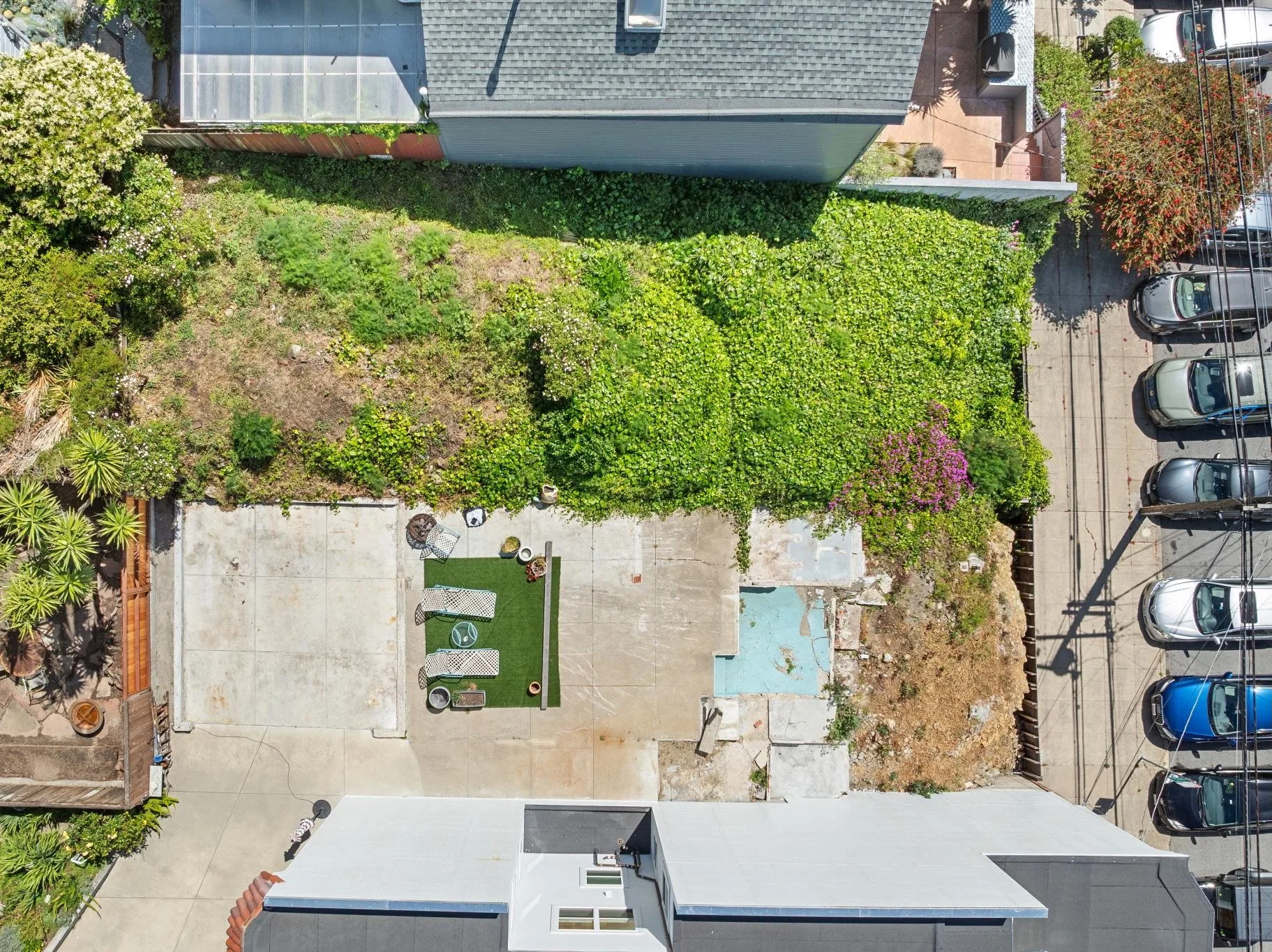 An aerial view of a backyard with a concrete patio, decorative furniture, a small artificial grass area, and a large garden with green plants and trees, adjacent to a parking lot with parked cars.