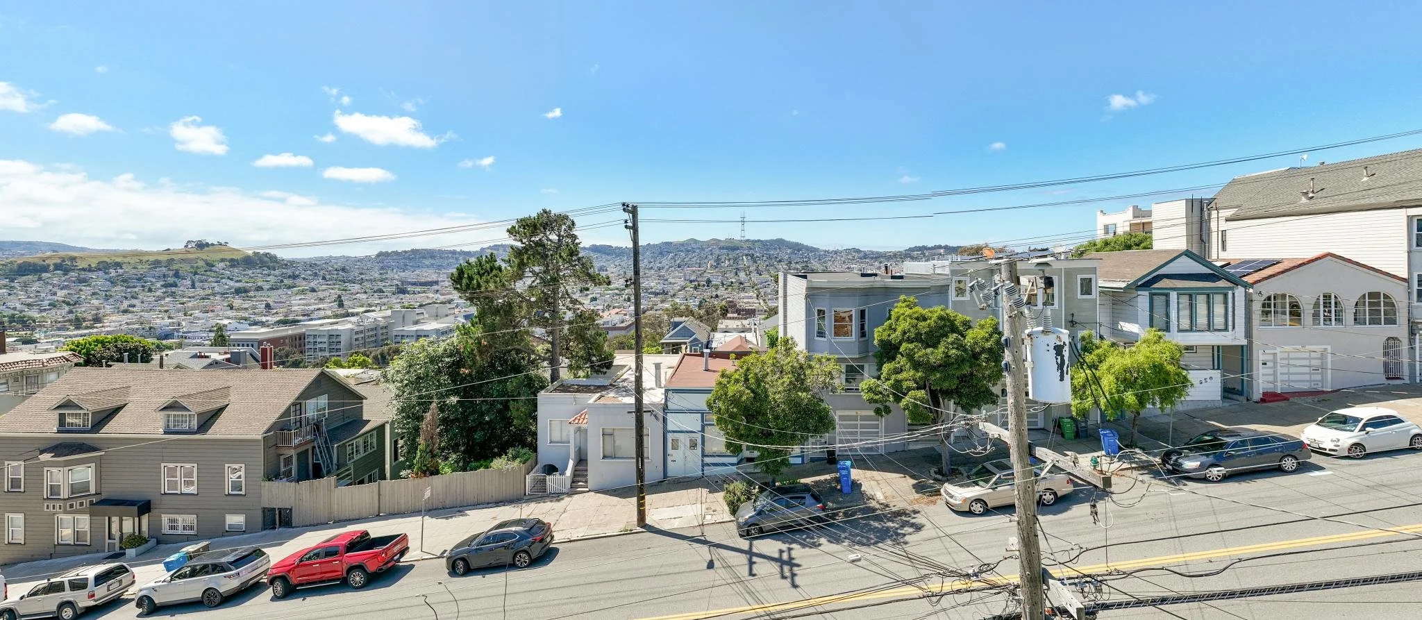 View of a neighborhood with houses, trees, parked cars, and utility poles, with a cityscape and rolling hills in the distance under a clear blue sky.