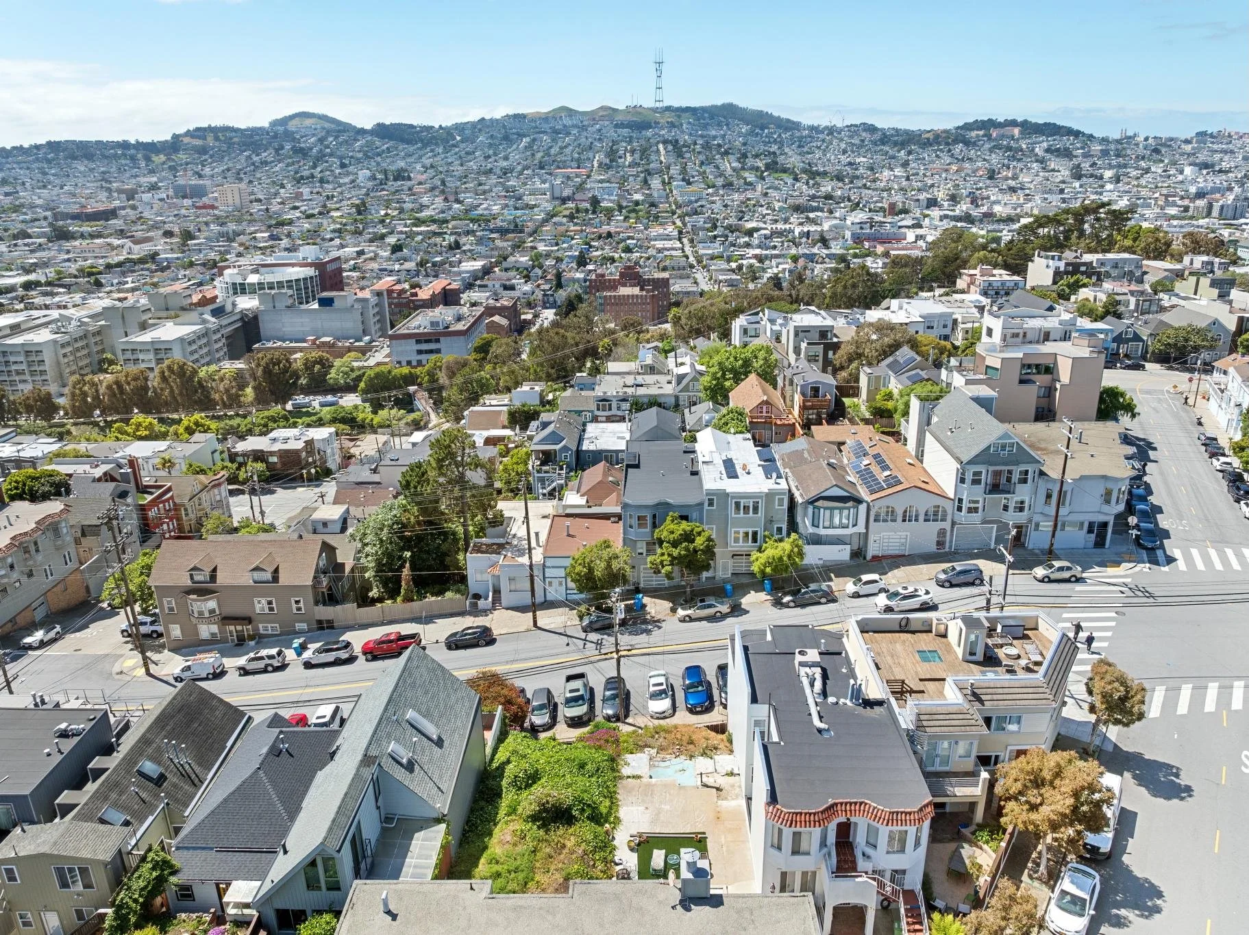 An aerial view of a dense urban neighborhood in San Francisco, with numerous houses, apartment buildings, and streets, showing a hilly landscape with some greenery and radio towers on the hilltop in the background.