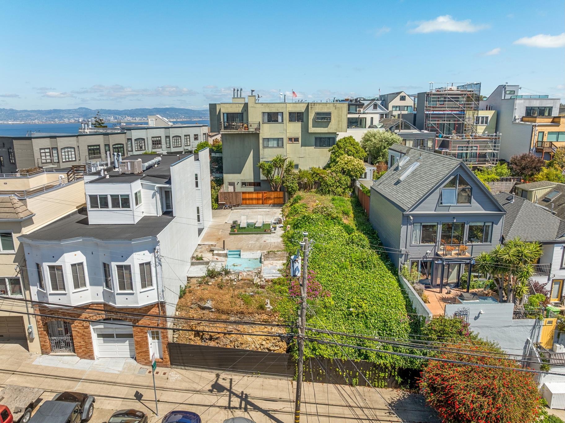 View of a neighborhood with various houses, some under construction, and a backyard with a small pool, surrounded by greenery and a fence. In the background, water and distant hills are visible under a partly cloudy sky.