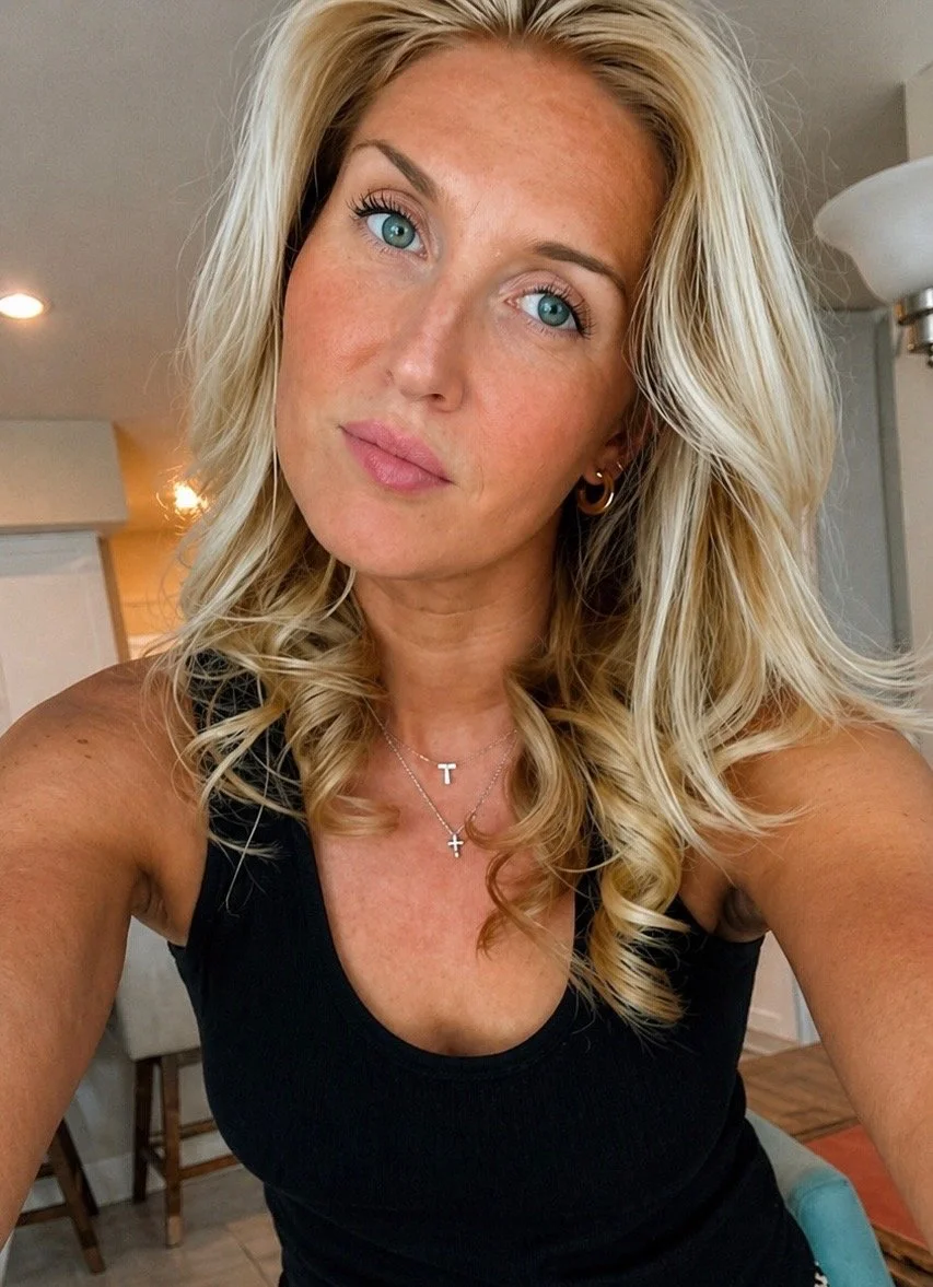 A woman with long blonde hair and blue eyes taking a selfie indoors, wearing a black top and layered necklaces.