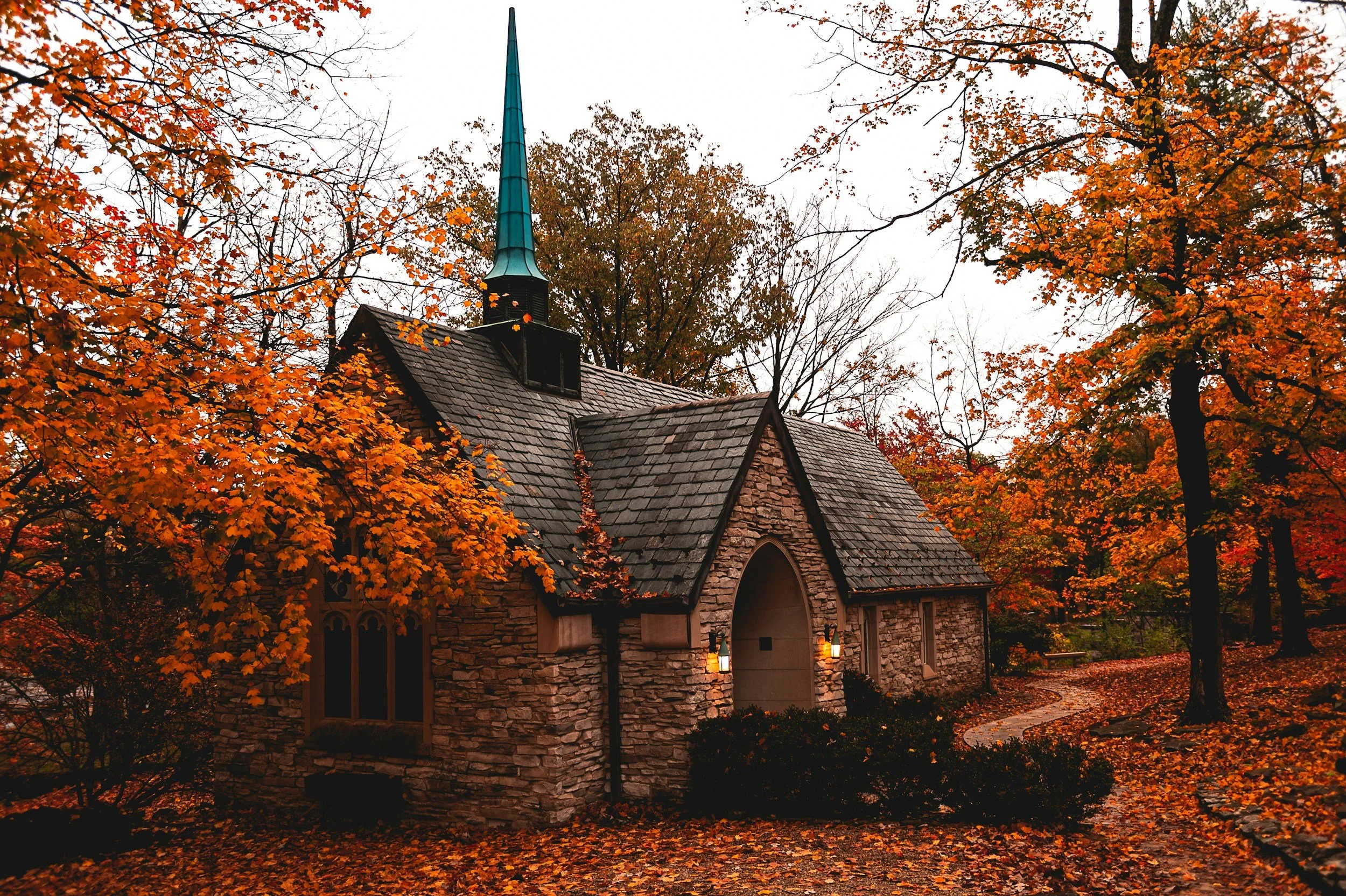 A stone church with a steep roof and a teal steeple, surrounded by fall foliage with orange and yellow leaves, overcast sky.