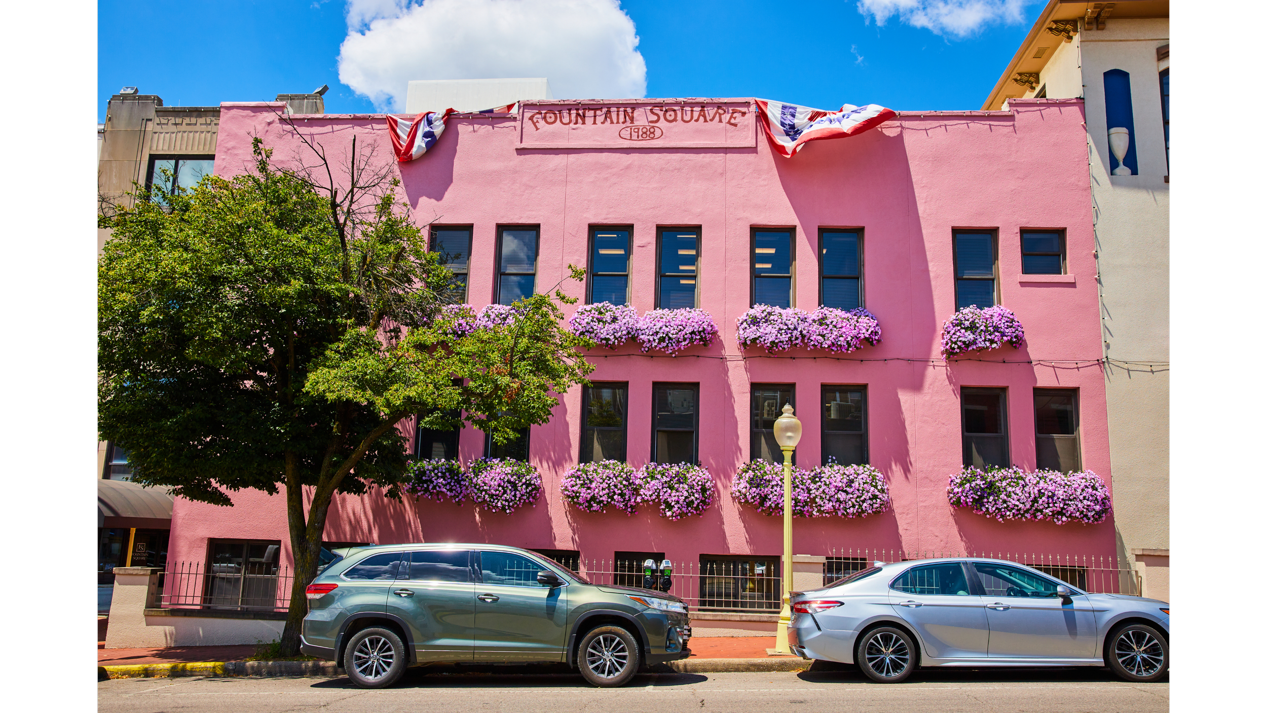 Pink building labeled 'Fountain Square 1888' with flower boxes, trees, parked cars, and a blue sky with clouds.