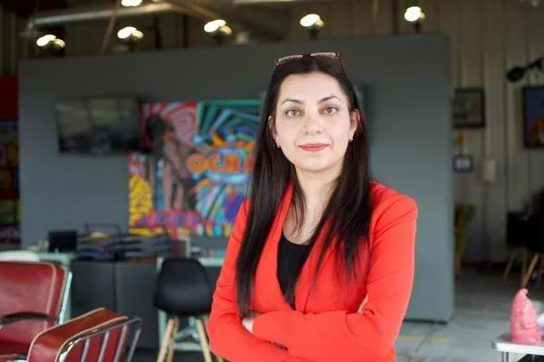 Woman with long dark hair wearing a red jacket and black top standing inside a modern indoor space with colorful wall art in the background.