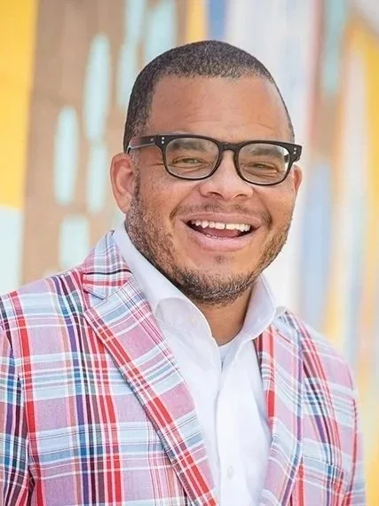 Donald Griffin Jr. pictured smiling with glasses, wearing a plaid blazer and white shirt, standing in colorful background.