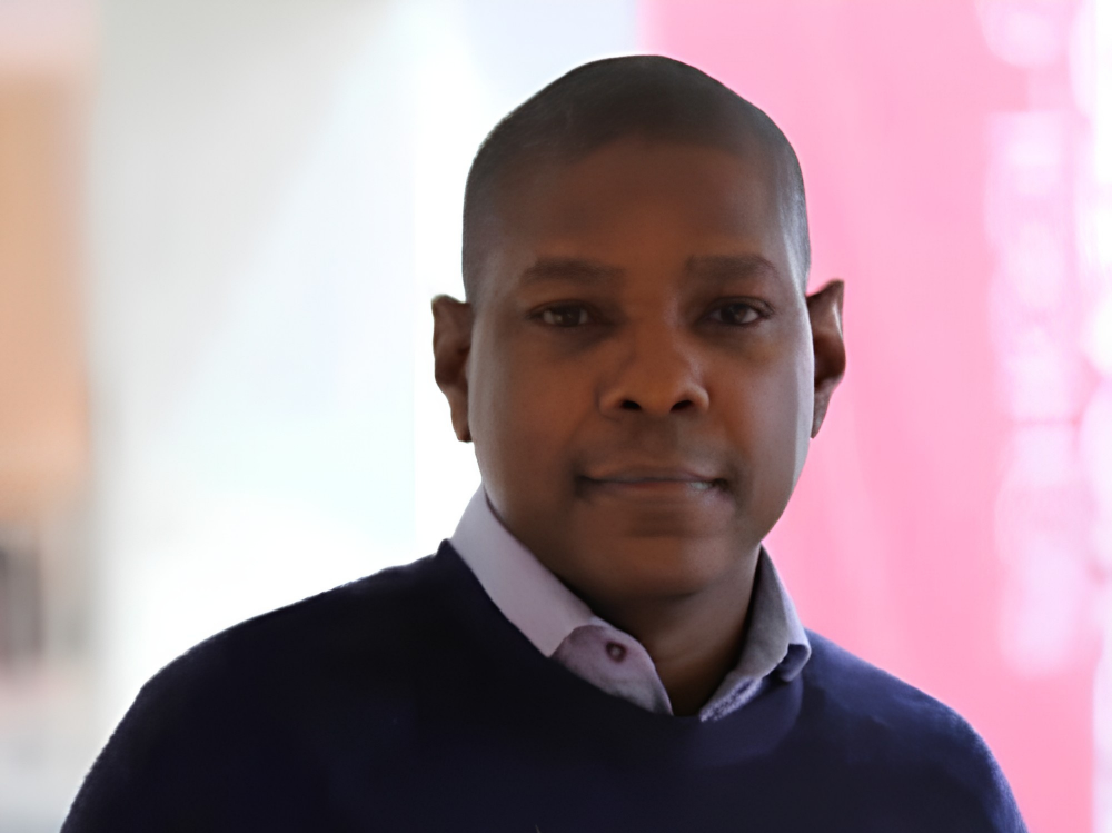 A man with short hair in a collared shirt and dark sweater, standing indoors in front of a pink wall.