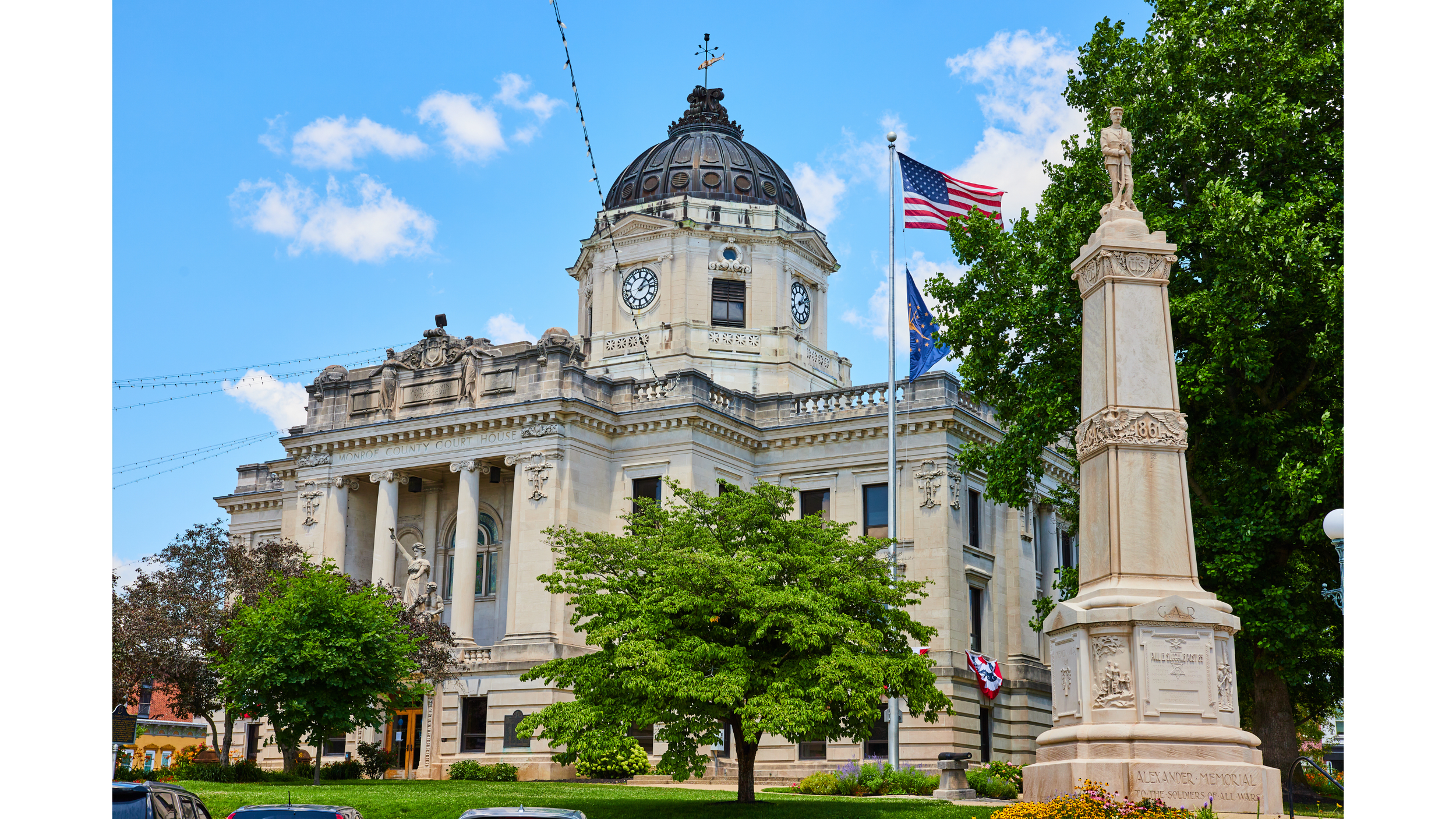 Historic Monroe County courthouse building with clock tower, American flag and Indiana state flag, statue on pedestal, lush green trees, bright blue sky with clouds.