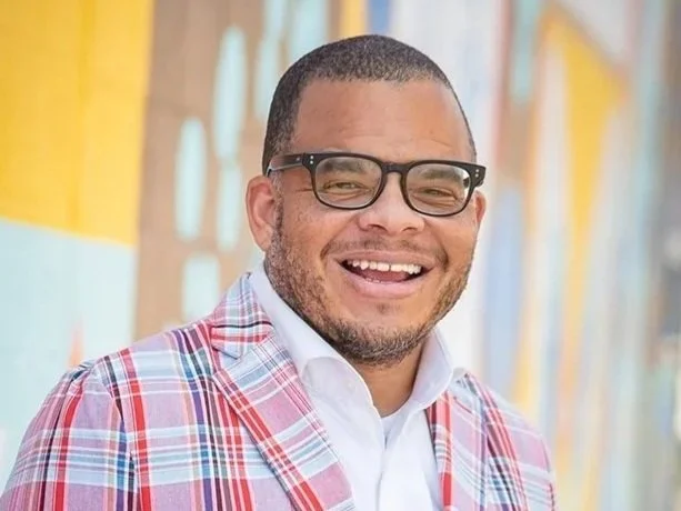 Donald Griffin Jr. pictured smiling with glasses, wearing a plaid blazer and white shirt, standing in colorful background.