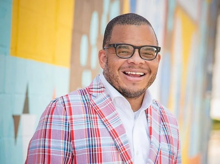 A smiling man with glasses wearing a pink plaid jacket and a white shirt, standing in front of a colorful mural.