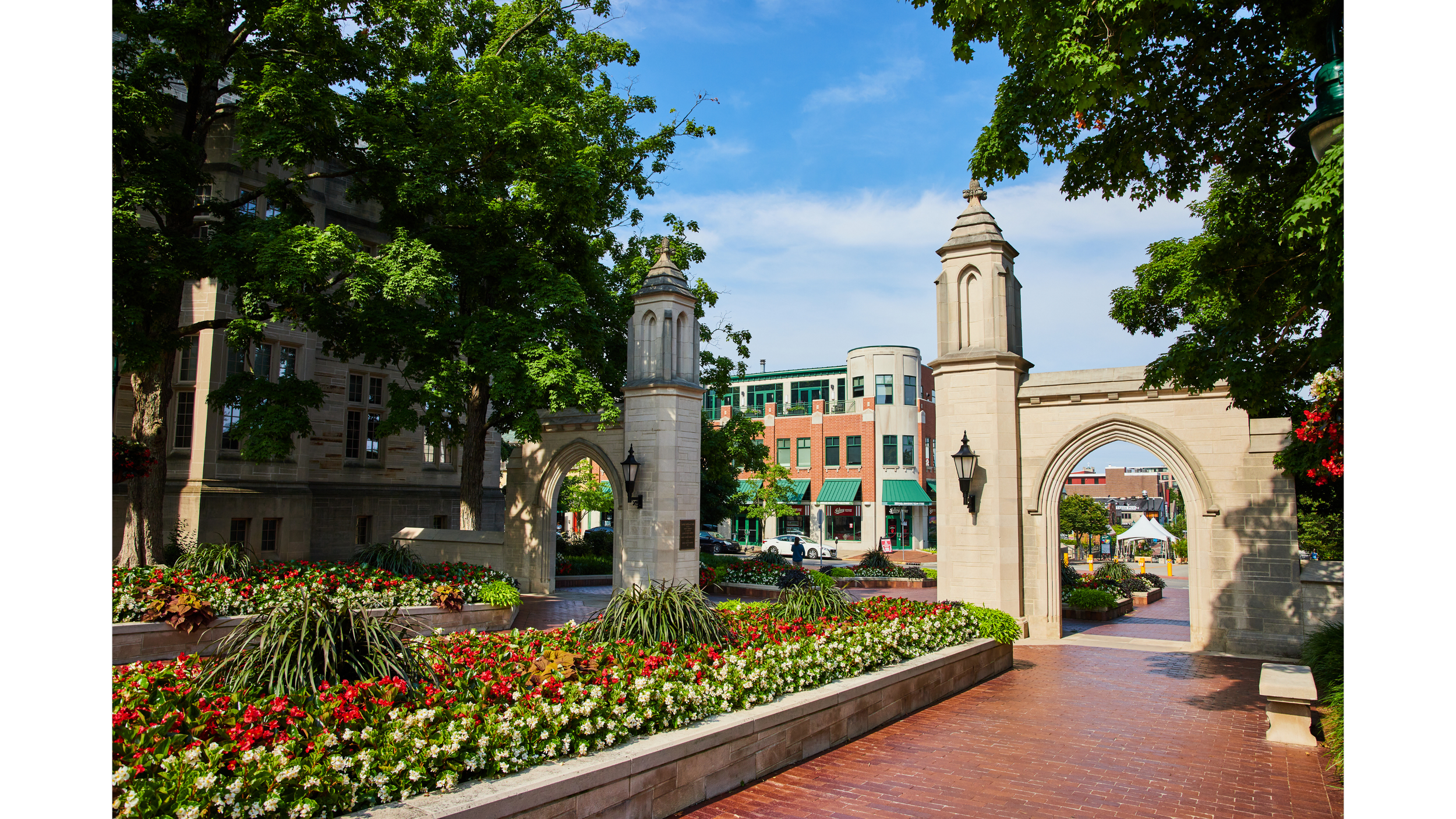 A historic stone gate with two towers, decorated arches, and a view of a modern building and street with shops and cars in the background, shaded by green trees and surrounded by colorful flowers.