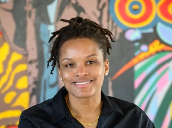 Woman with dreadlocks smiling, wearing a black shirt and a gold necklace, standing in front of a colorful mural.