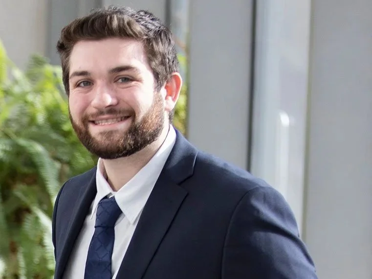 A young man in a navy suit, white shirt, and navy tie smiling in front of a window and greenery.