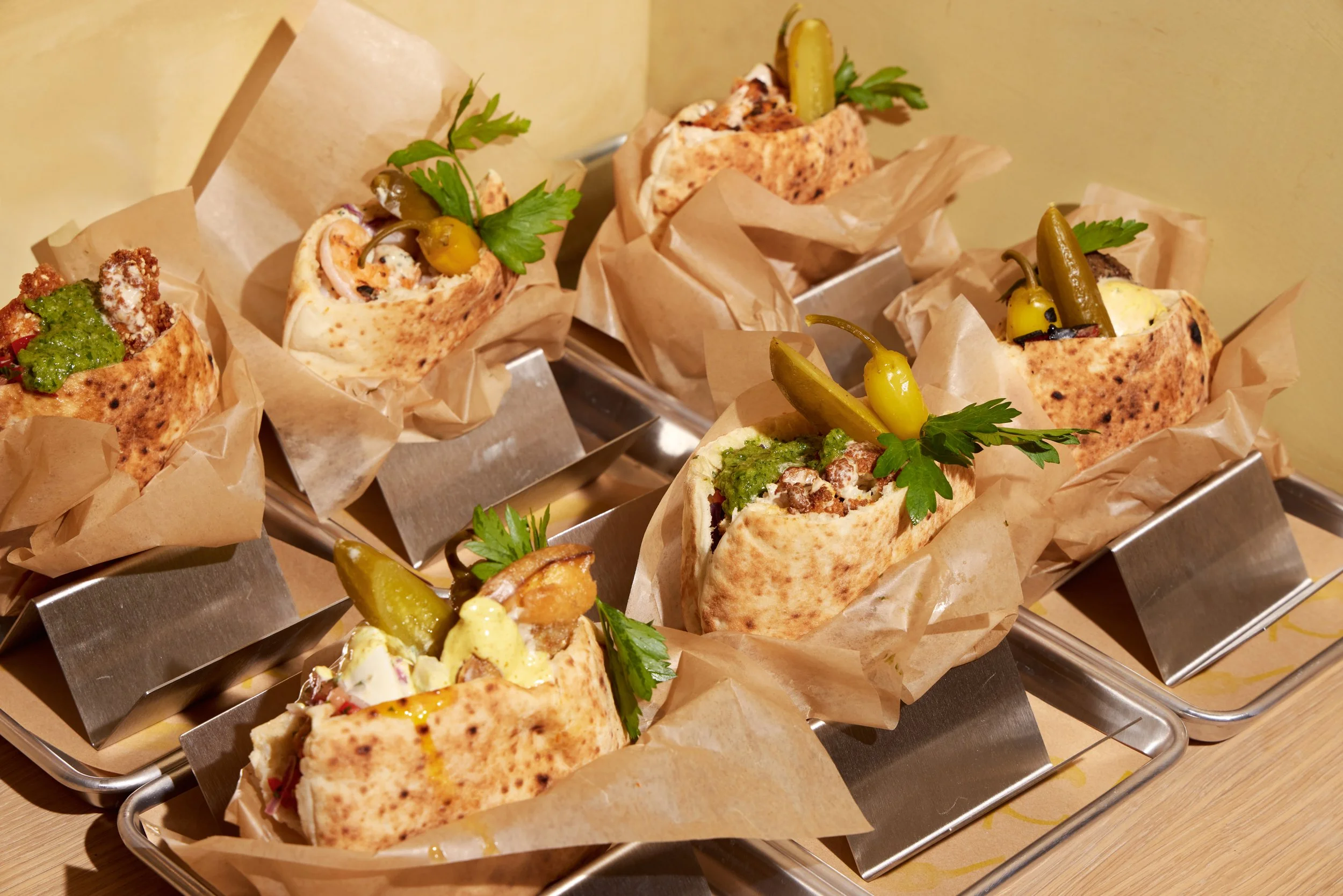 Pita sandwiches of different types wrapped in paper in metal trays on a table.