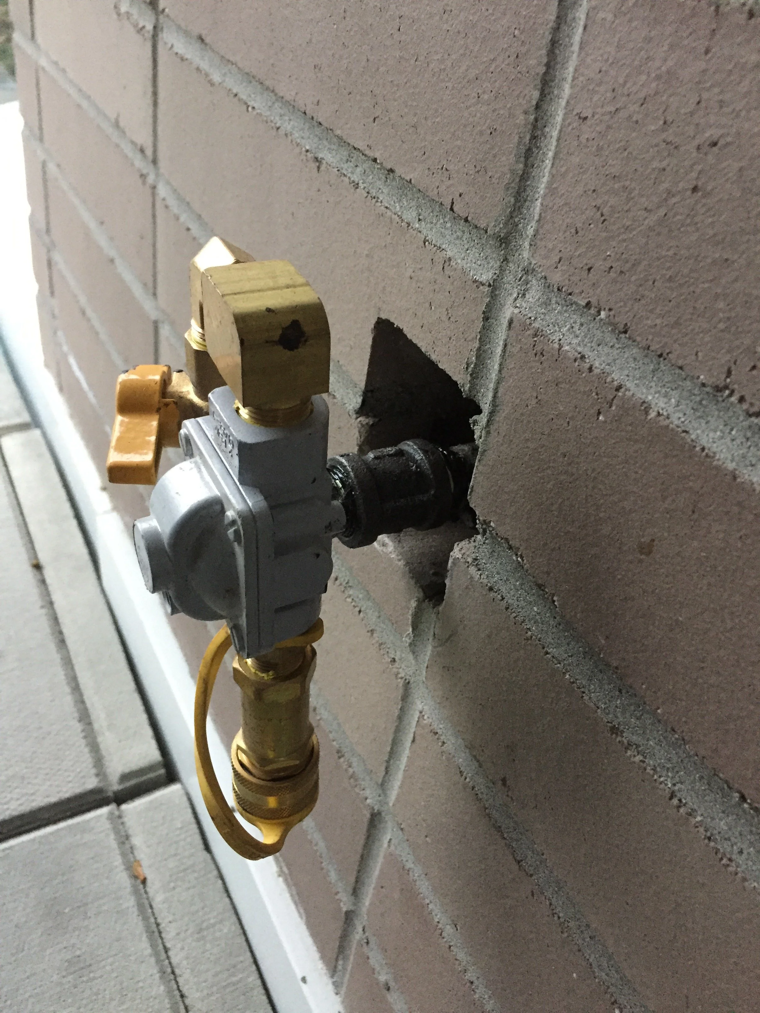 A close-up of a plumbing connection on a brick wall, showing a pipe fitting BBQ box with a gray control valve and brass fittings.