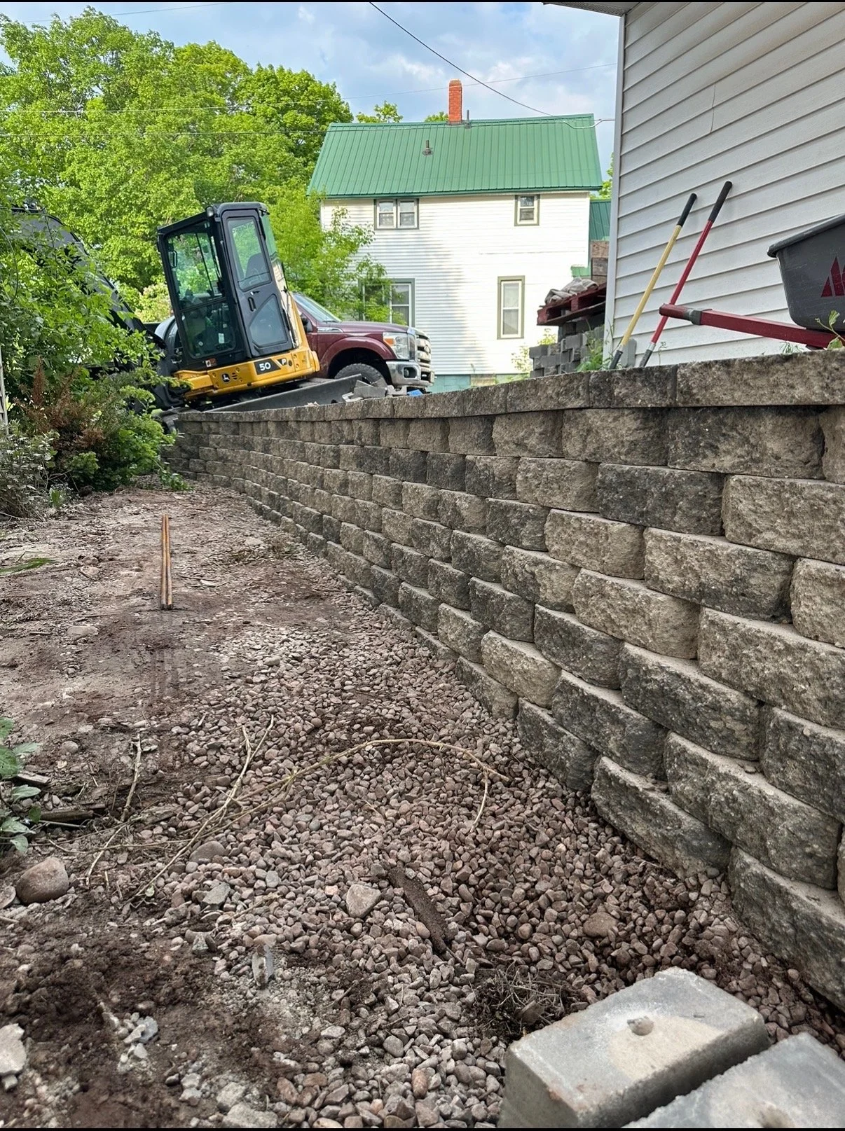 A flower bed with a retaining wall built with stone blocks. Construction tools and a mini excavator are present, with a house and trees in the background.