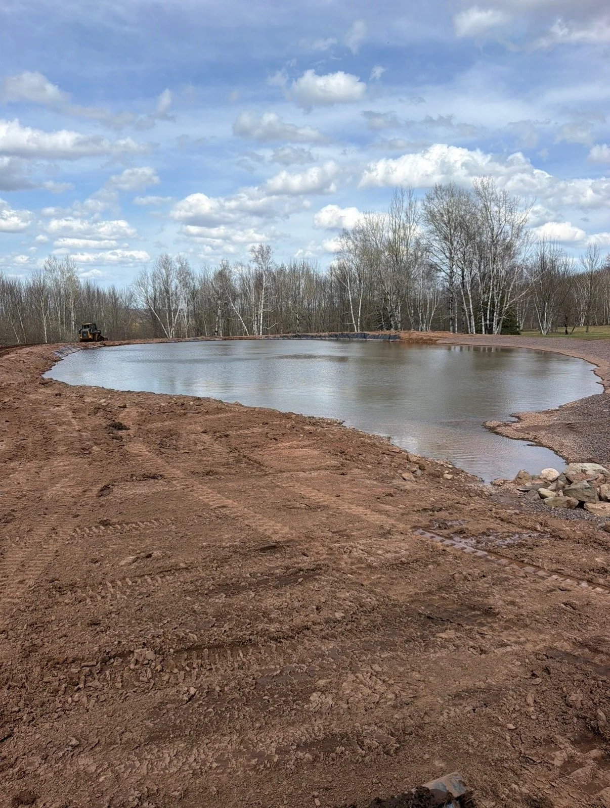 Construction site with a small pond, dirt ground, and trees in the background under a partly cloudy sky.
