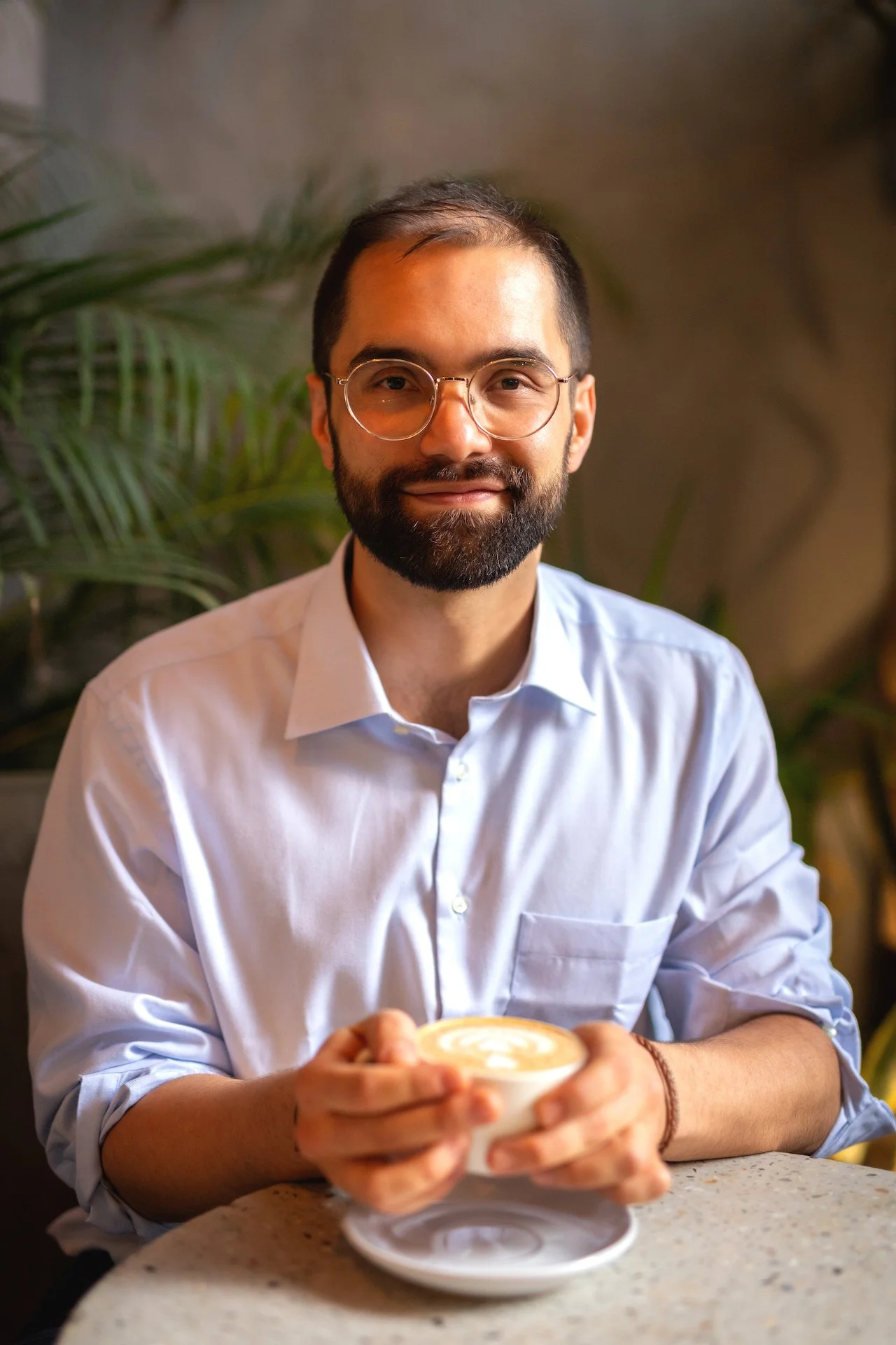 A man with glasses and a beard wearing a white shirt, sitting at a table and holding a cup of coffee or latte with foam art.